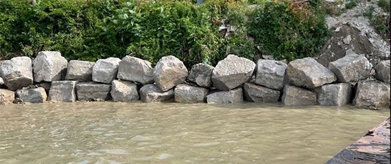 A row of large, stacked grey boulders acting as a shoreline barrier against murky, light-brown water and green bushes.