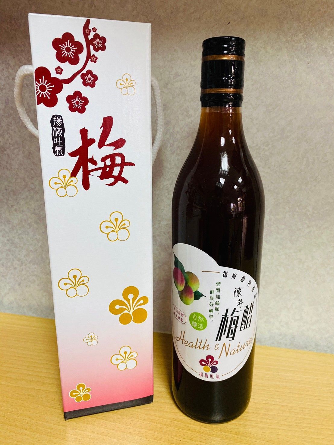 一瓶深色液體，貼有白色標籤，旁邊是一個配套的禮盒，兩者都印有日文字和梅花圖案。