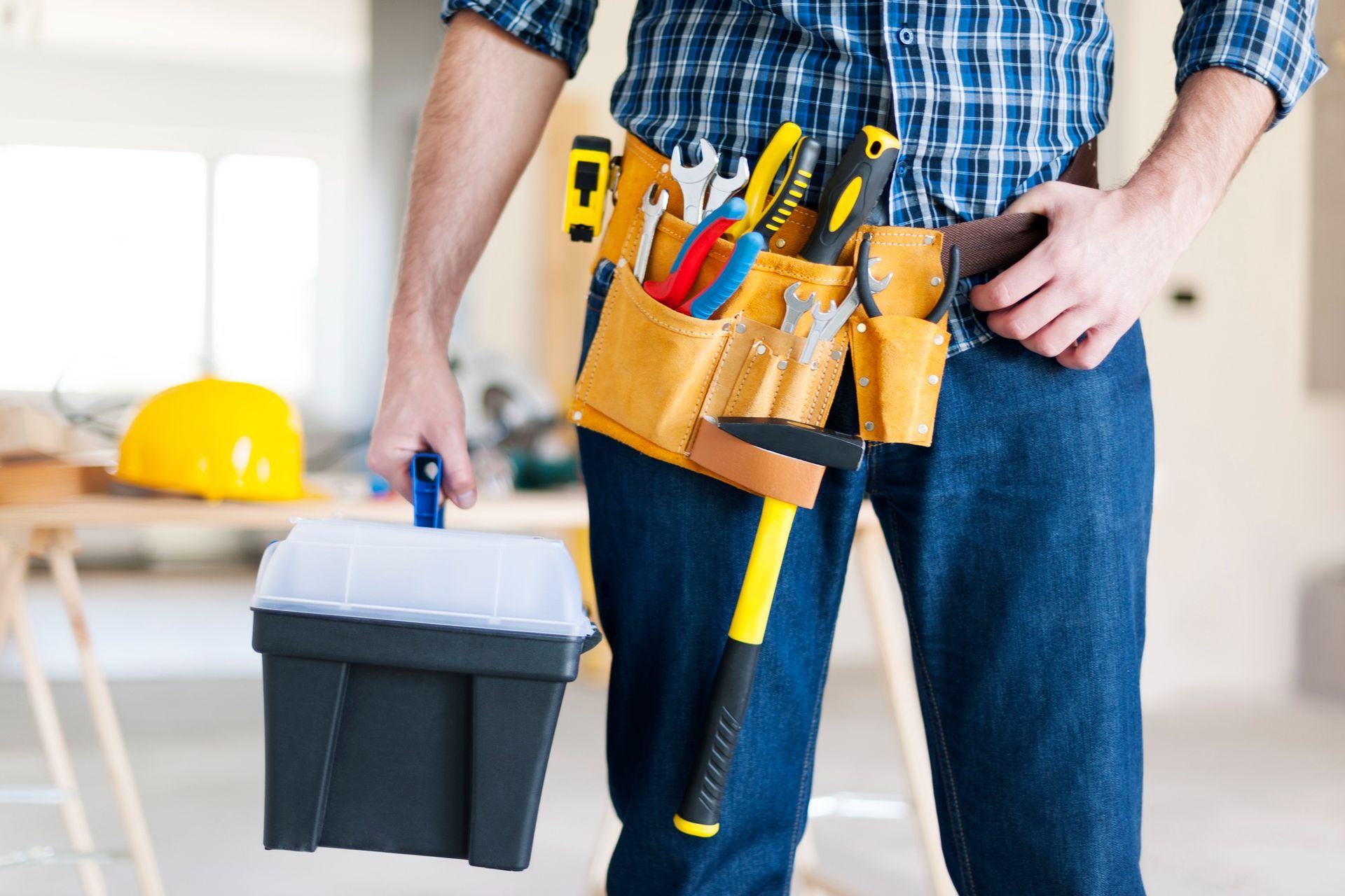 A worker in a plaid shirt and tool belt holds a paint tray in a construction setting with a yellow hard hat nearby.