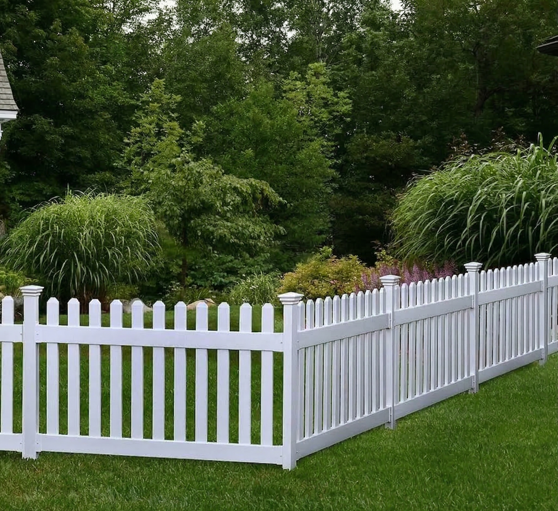 A white picket fence stands in a lush green backyard, bordered by trees and ornamental grasses.