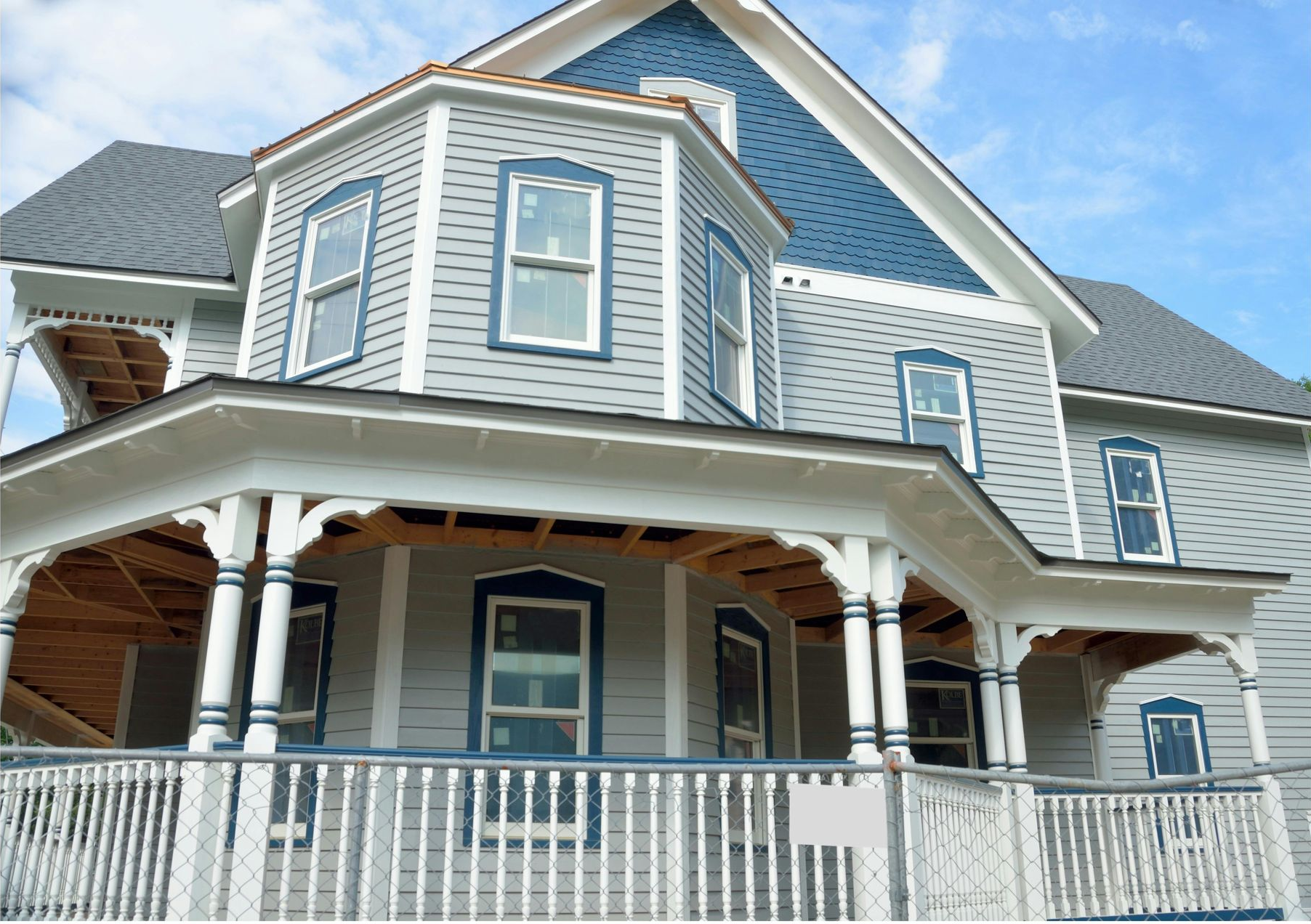 A two-story grey Victorian house with white trim, a wrap-around porch, and blue decorative siding on the front gable.