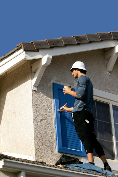 A person wearing a white hat and dark clothes stands on a roof, painting a blue window shutter.