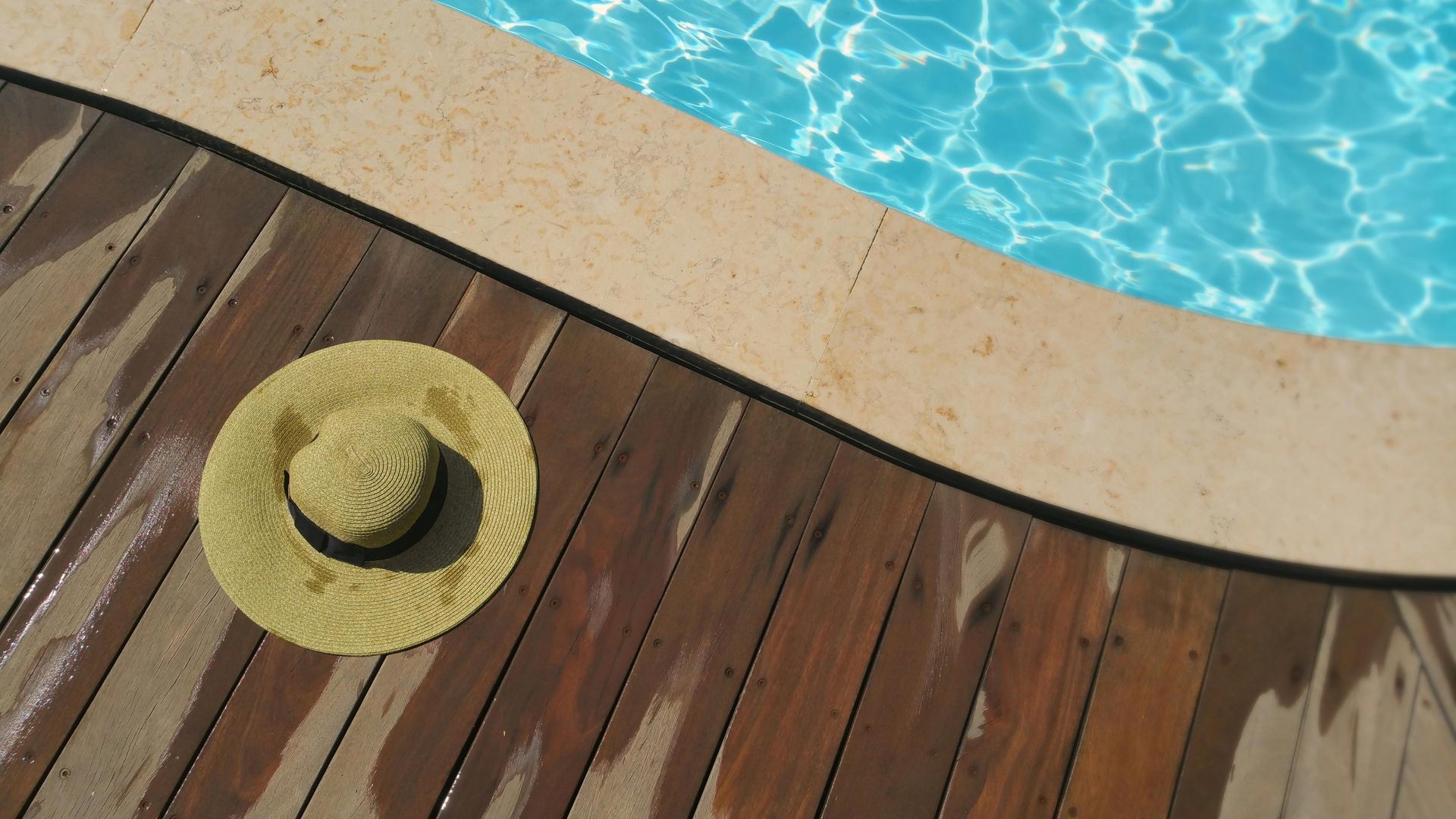 A light-colored straw sun hat rests on a wooden pool deck next to the edge of a swimming pool with blue, rippling water.