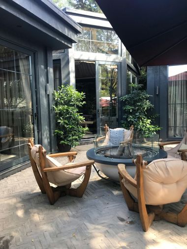 A patio with two wooden chairs, beige cushions, and a fire pit, set against a dark grey building with glass doors.