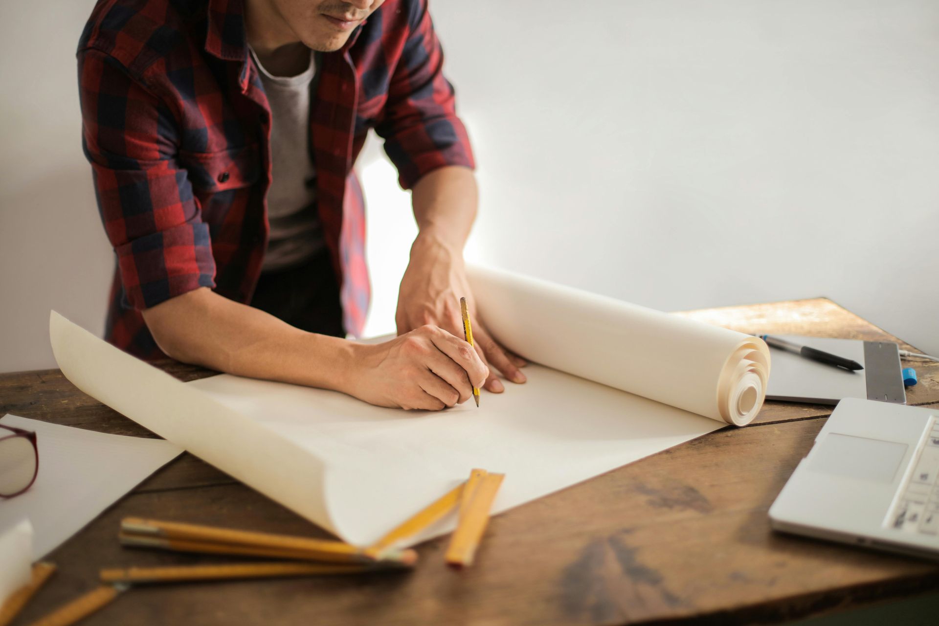 Person in a red plaid shirt drawing with a pencil on a large roll of paper spread across a wooden desk.