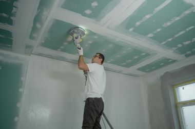 A person stands on a ladder, using a power sander to smooth the seams of green drywall panels on a ceiling.