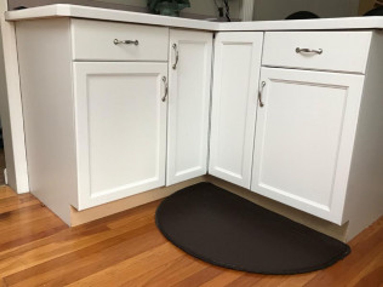 White kitchen cabinets in a corner with a dark brown, half-moon shaped floor mat on a wood floor.