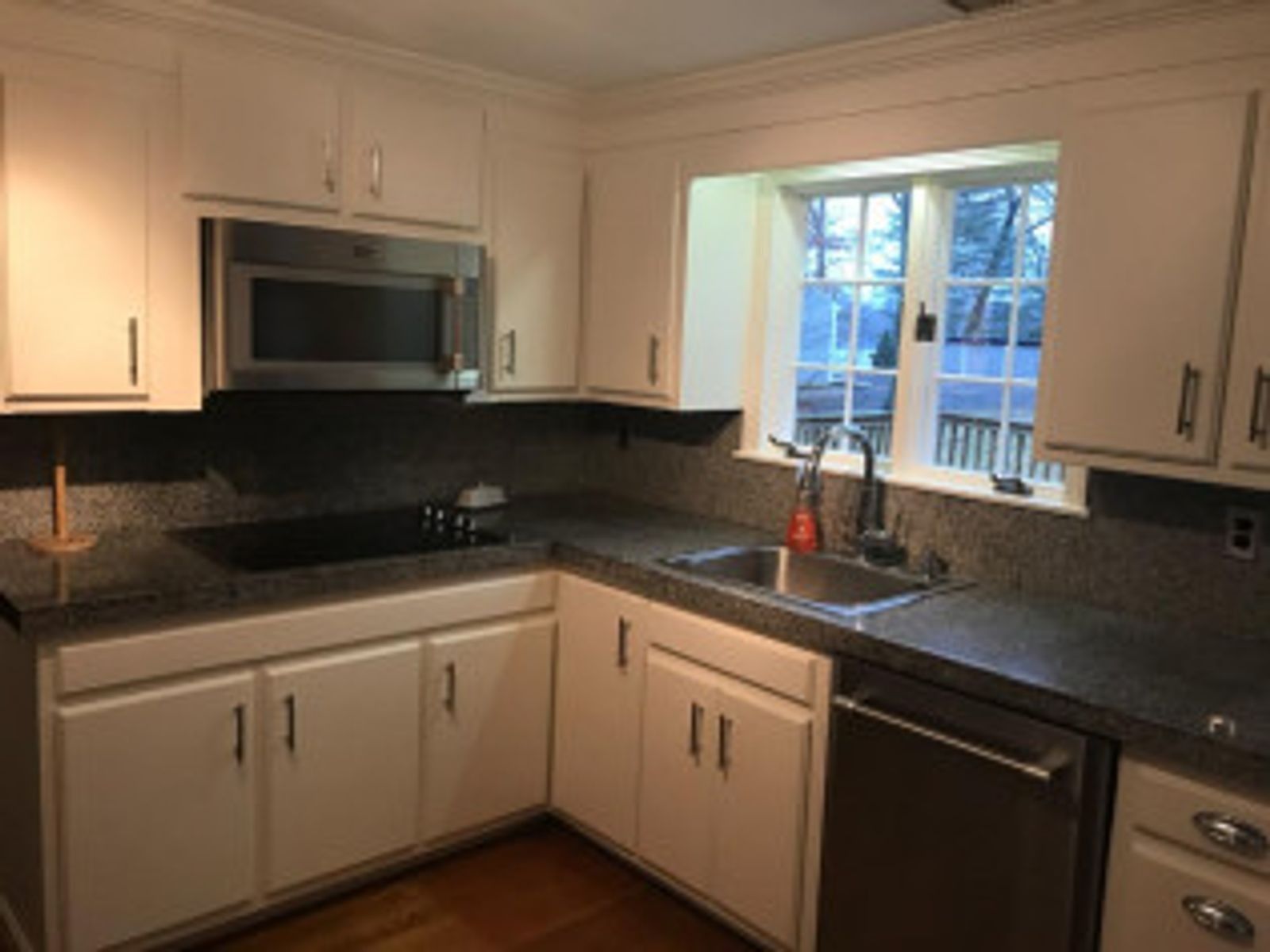 White kitchen with stainless steel appliances, dark countertops, and a window.