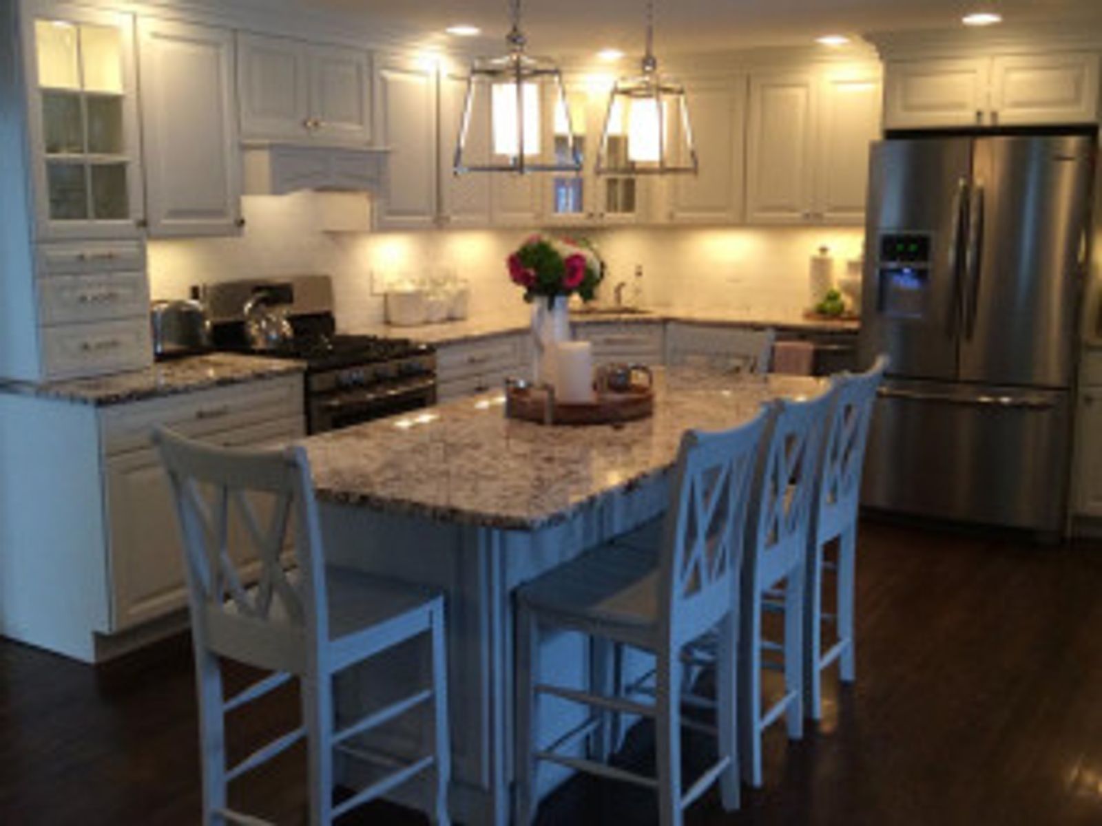 White kitchen with island, granite countertop, bar stools, stainless steel fridge, and pendant lights.