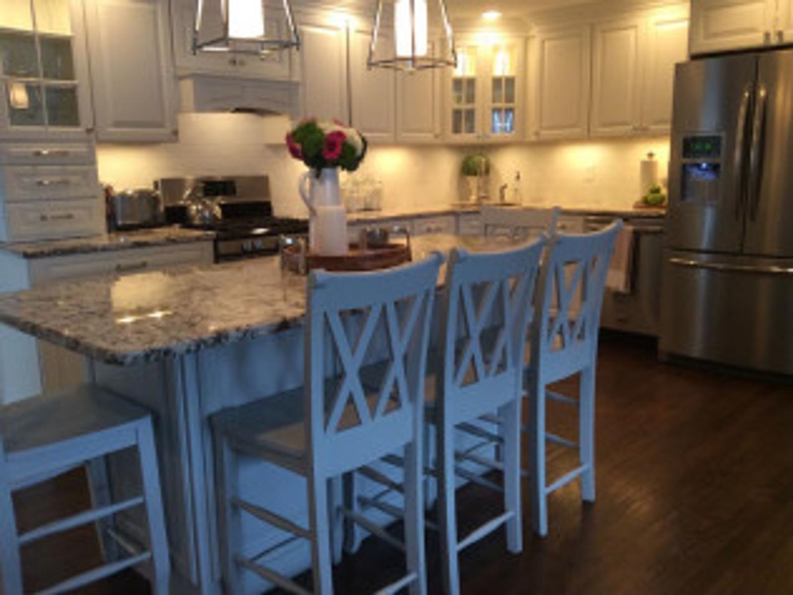 A bright white kitchen with a granite island, stools, and stainless steel refrigerator.