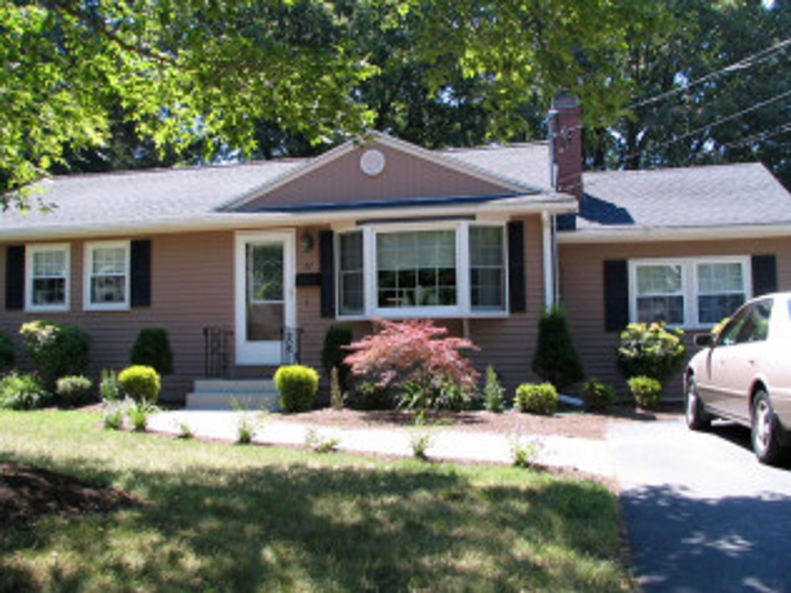 Tan ranch house with dark shutters, front yard landscaping, driveway, and a car parked on it.