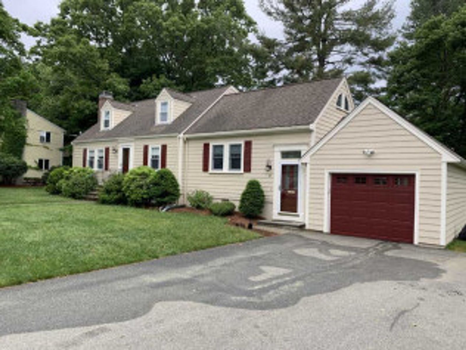 Beige house with a red garage door and shutters, green lawn and trees.