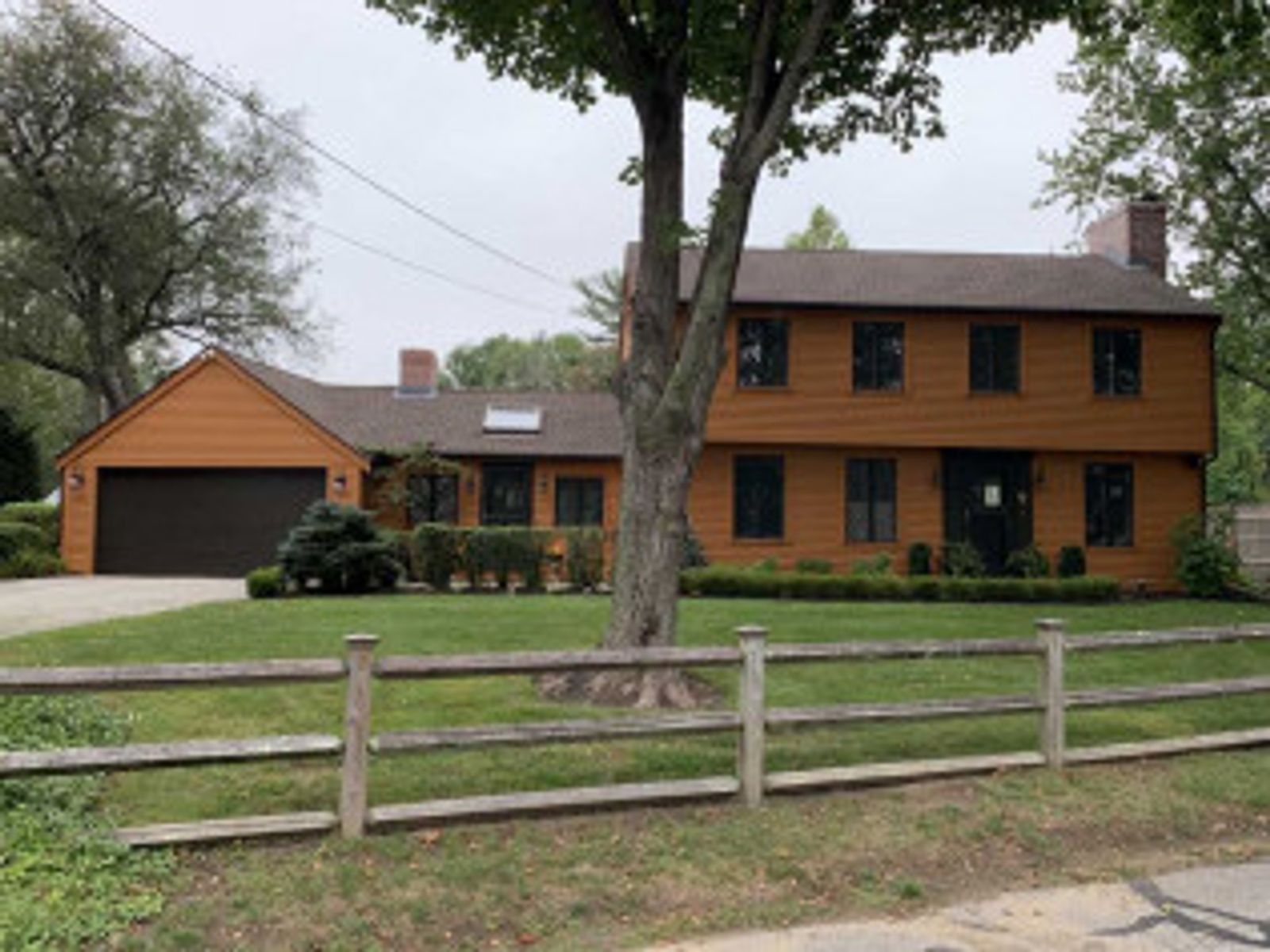 Brown two-story house with a wooden fence and a large tree in front. Green grass and overcast sky.