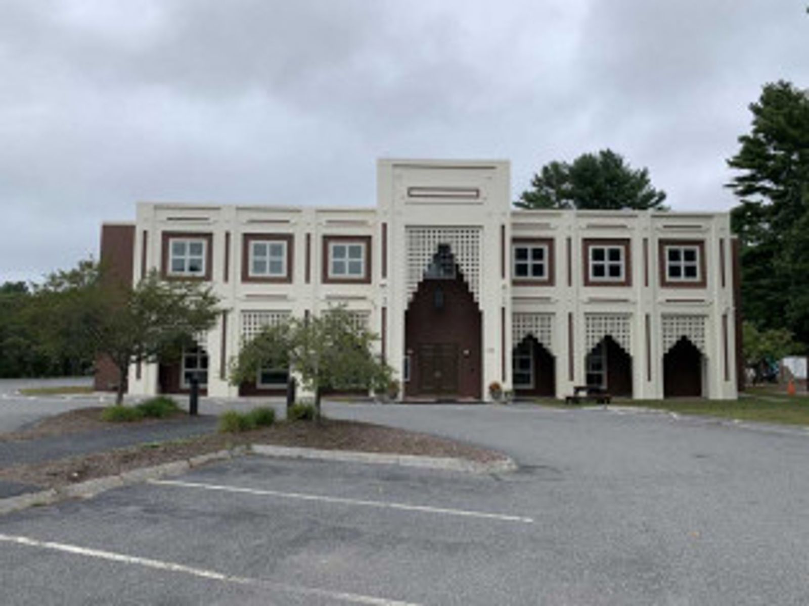 Beige and brown building with arched doorways and square windows, set on a parking lot on an overcast day.