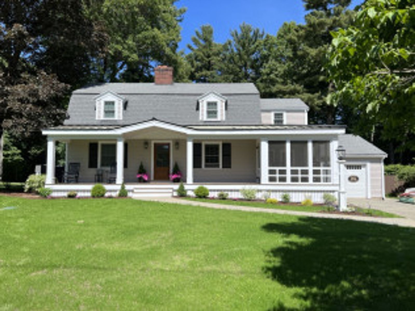 Beige Cape Cod home with porch, lawn, and green trees under a bright blue sky.
