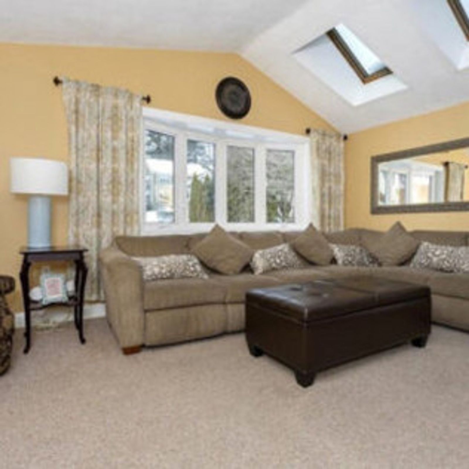 Cozy living room with beige sofa, brown ottoman, and large windows. Skylights and yellow walls provide natural light.