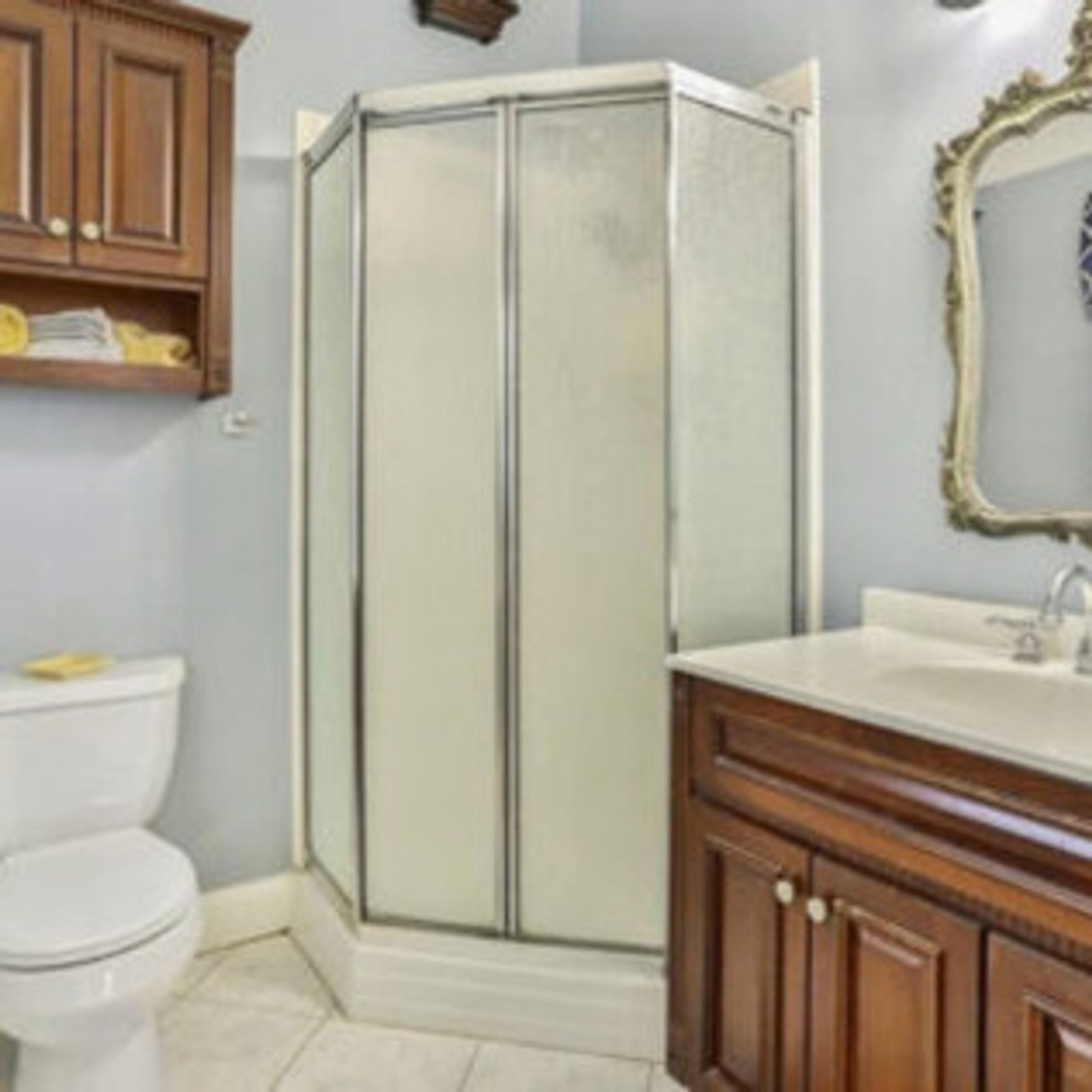 Bathroom with shower, vanity, toilet, and wooden cabinets. Light blue walls, white fixtures, and a decorative mirror.
