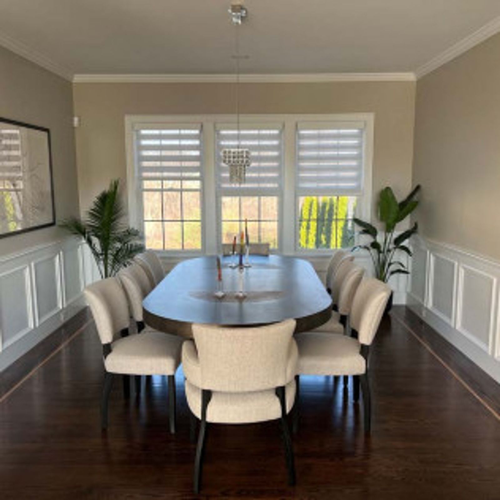 Dining room with oval table, cream chairs, large windows, and plants.