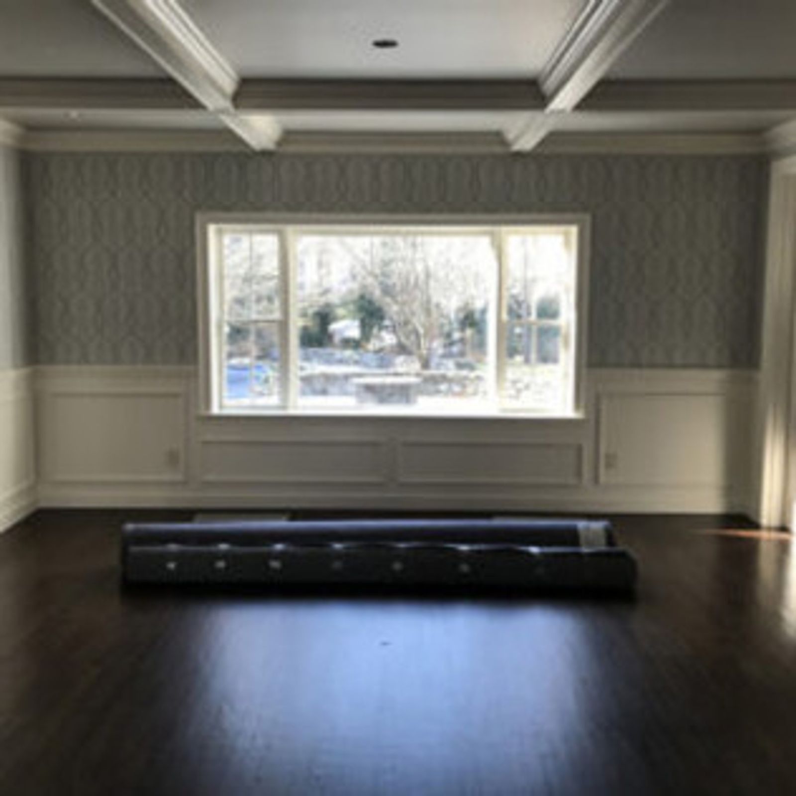 Empty room with dark wood floor, large window, and rolled-up rugs.