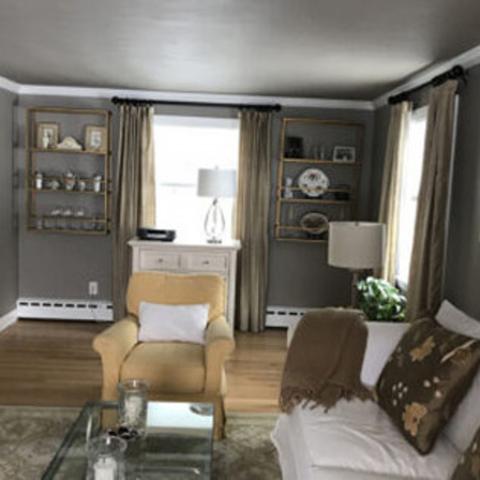 Living room with gold shelves, yellow armchair, white couch, beige curtains, and wooden floor.