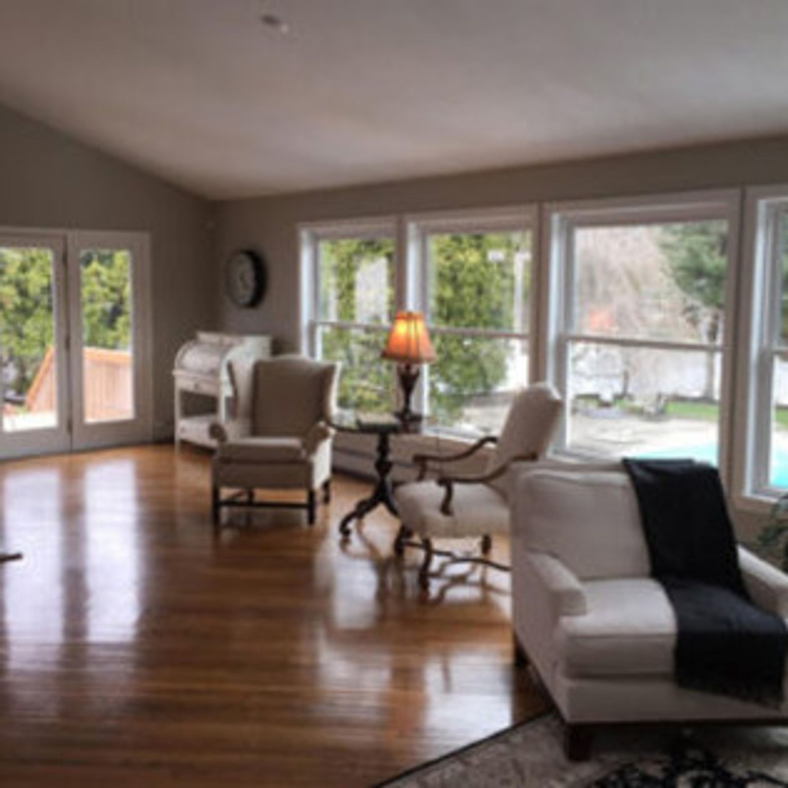 Living room with hardwood floors, white armchairs, and large windows overlooking a backyard.