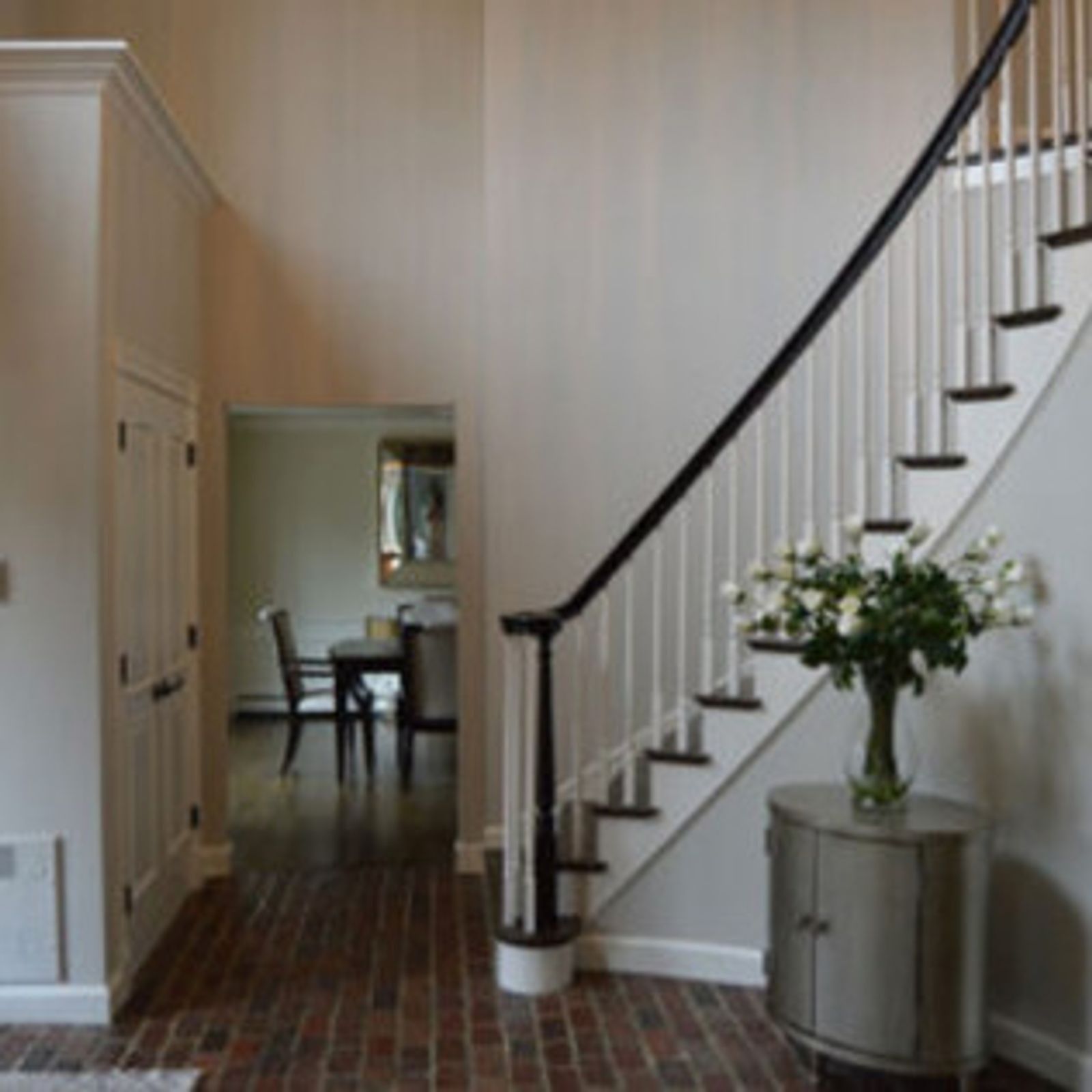 Entryway with stairs, brick floor, white walls, black railing. Table with flowers. Door to dining room.
