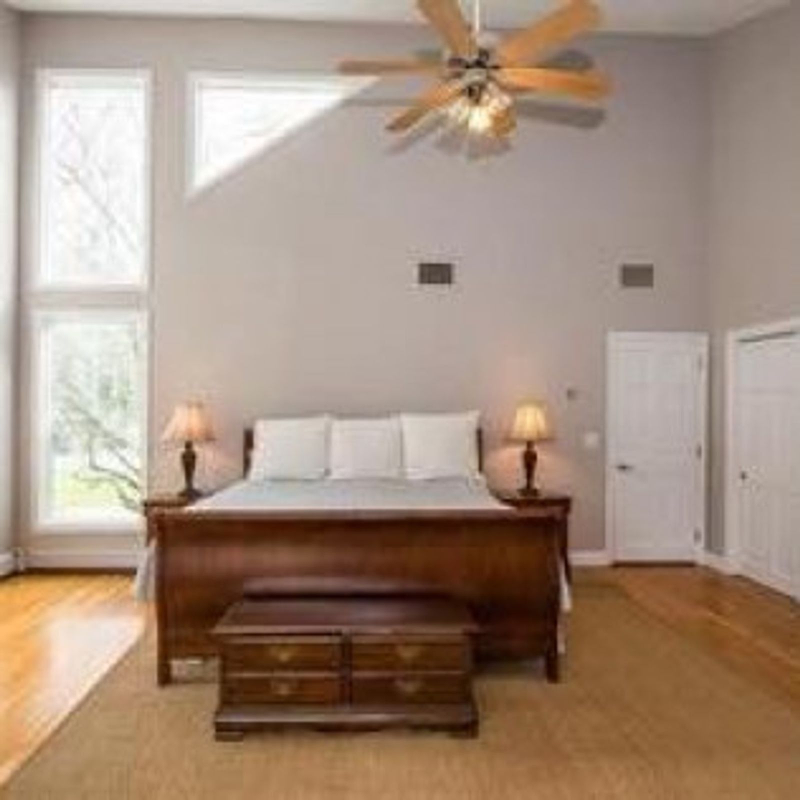 Bedroom with a bed, bench, ceiling fan, and large windows. The walls are light grey, and the floors are hardwood.