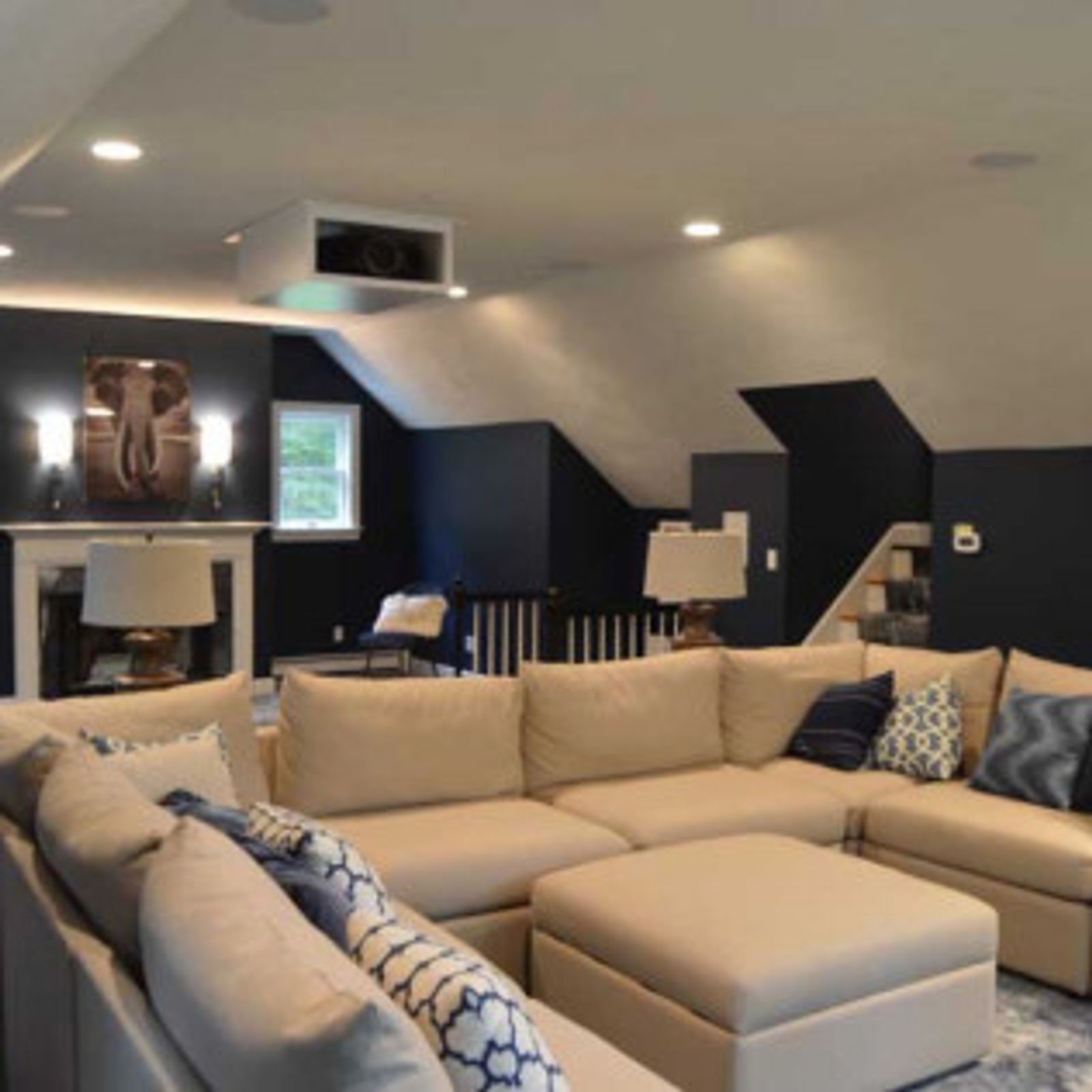 A cozy home theater with a large sectional sofa, fireplace, and projector screen. Dark blue walls and tan furniture.