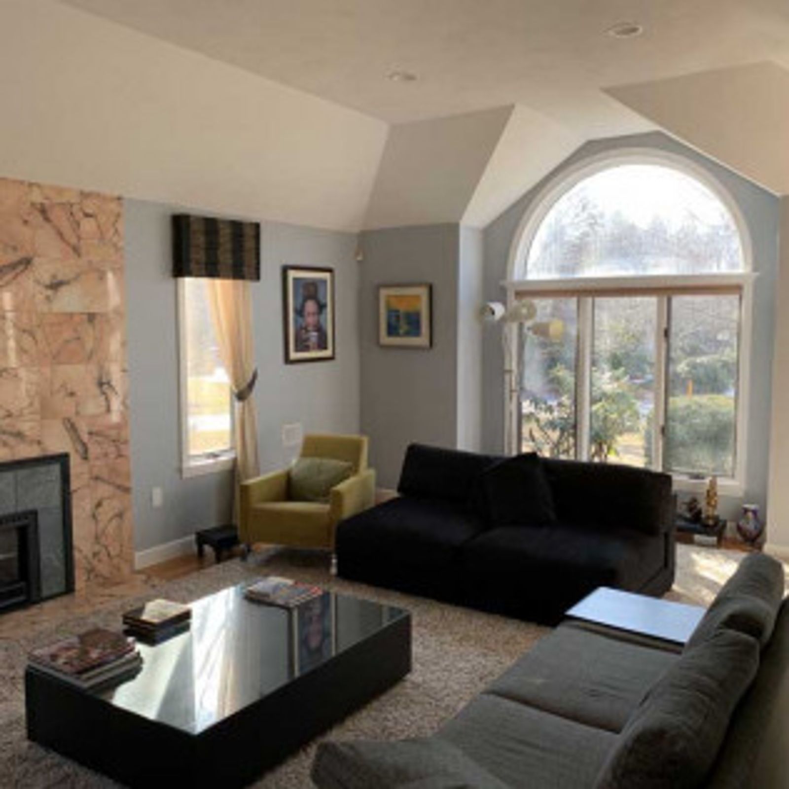 Living room with fireplace, sofas, and arched window. Beige walls, neutral-toned furniture, and sunlight.