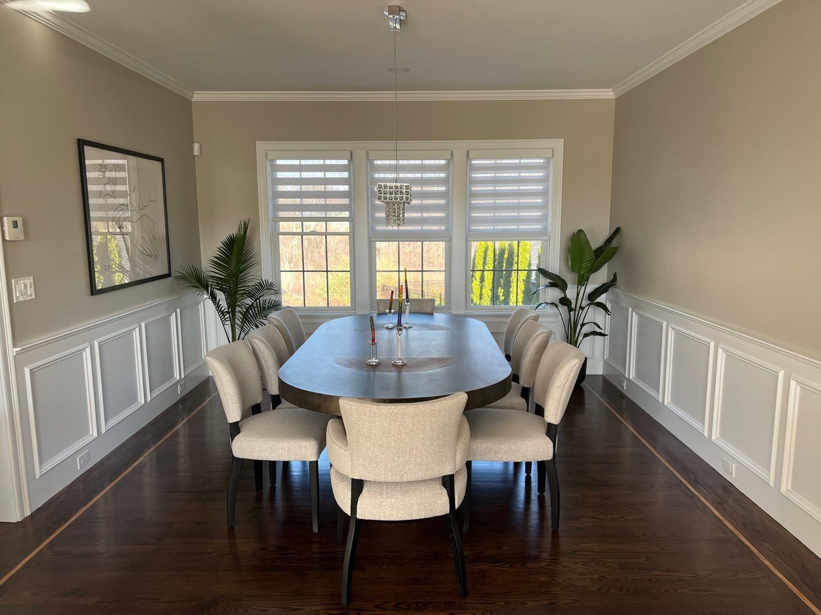 Dining room with an oval table and ten upholstered chairs. Windows with shades, and wainscoting on walls.