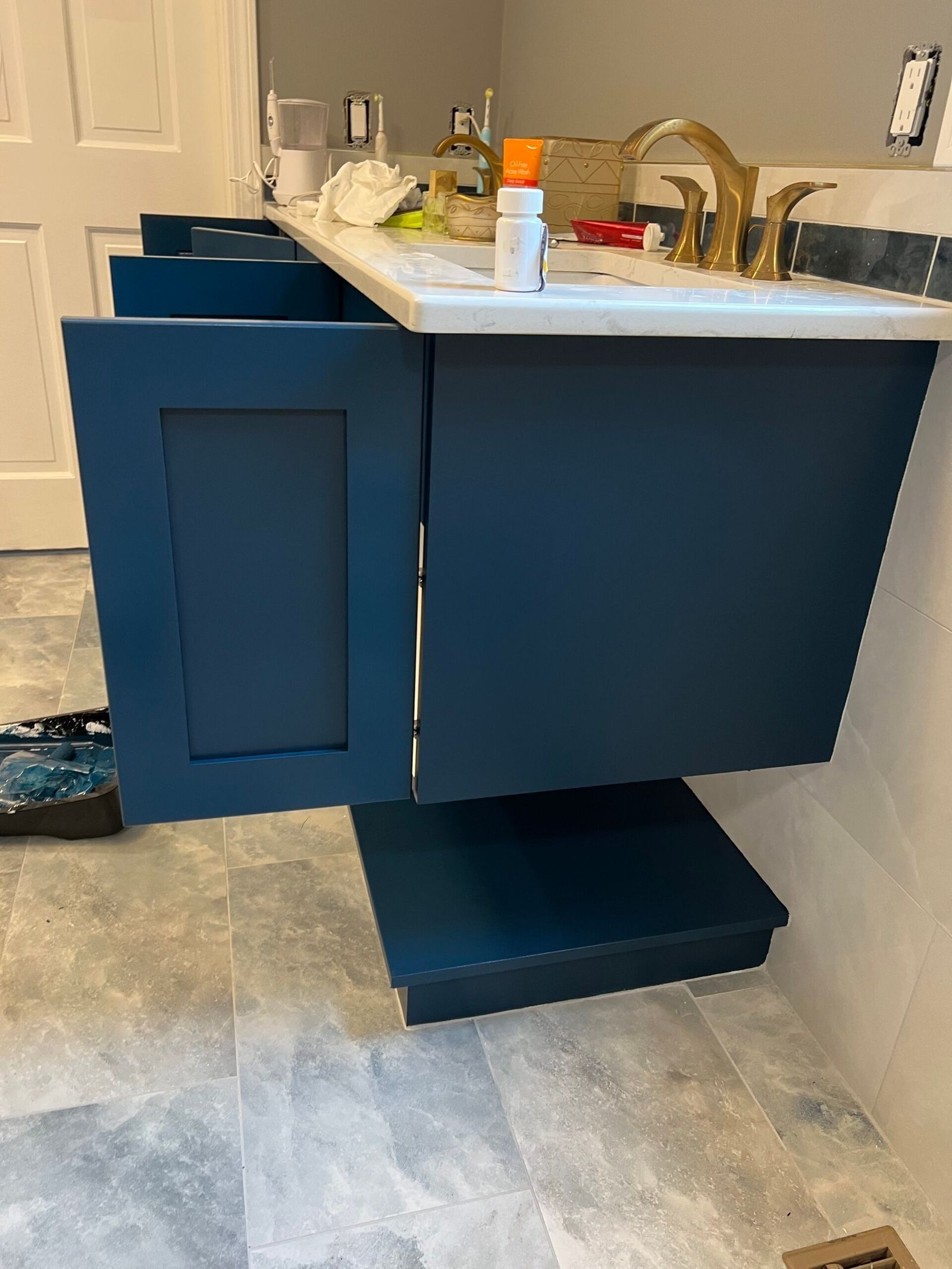 Navy blue bathroom vanity with an open door and a step stool, against a gray tiled floor.