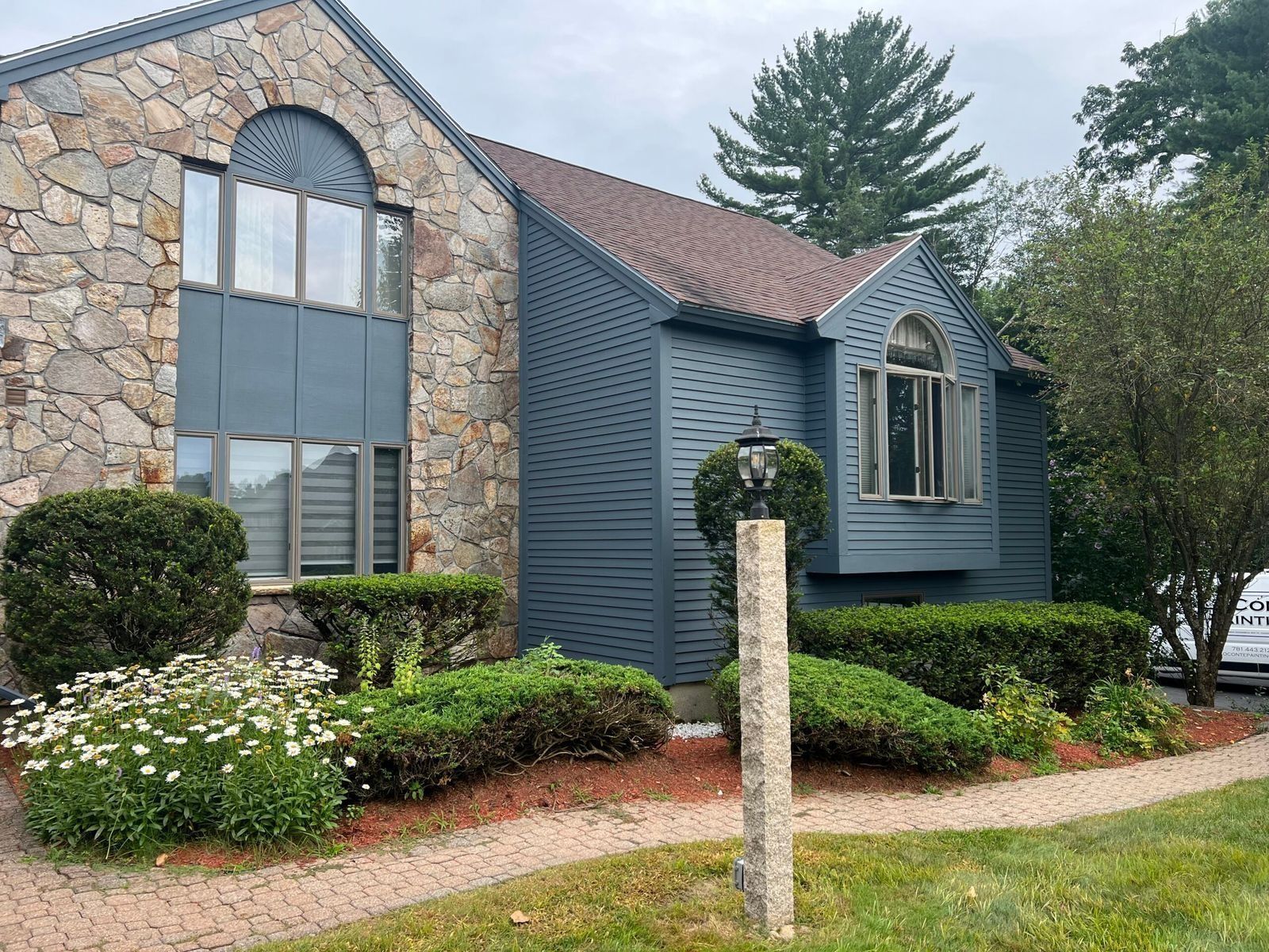 Stone and blue siding house with arched windows and brown roof, surrounded by bushes and a small lawn.