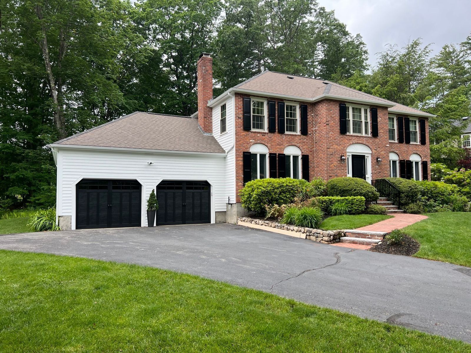 Two-story brick house with black shutters, attached two-car garage, and asphalt driveway. Green lawn and trees surround the home.
