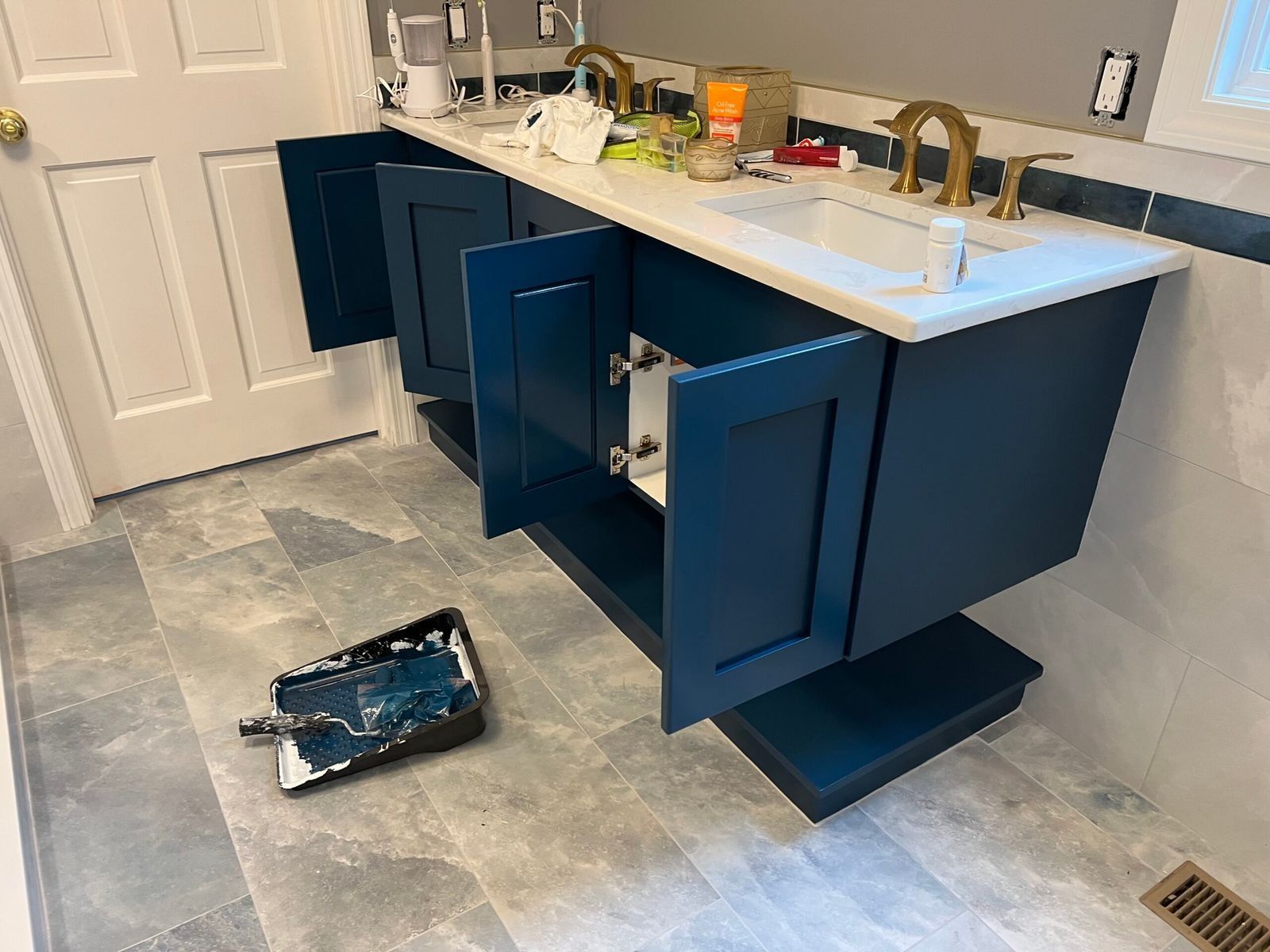 Blue vanity with open doors, gold fixtures, and paint supplies on tiled floor.