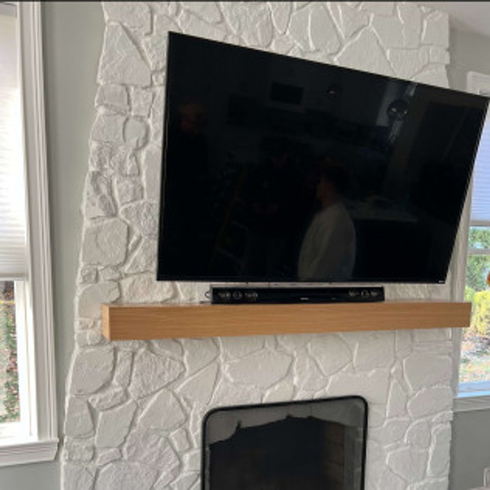 A TV mounted on a white stone fireplace with a wooden mantle and window blinds.