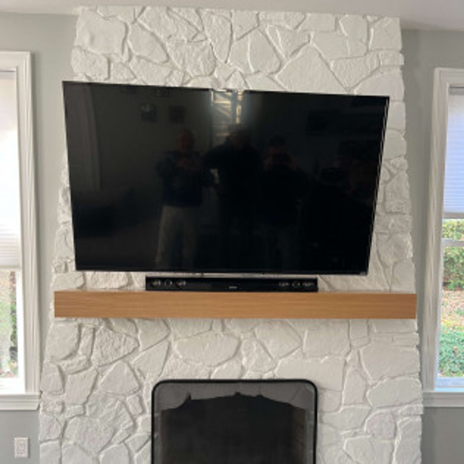 Large TV mounted on a white stone fireplace with a wooden shelf below. Windows are on either side.
