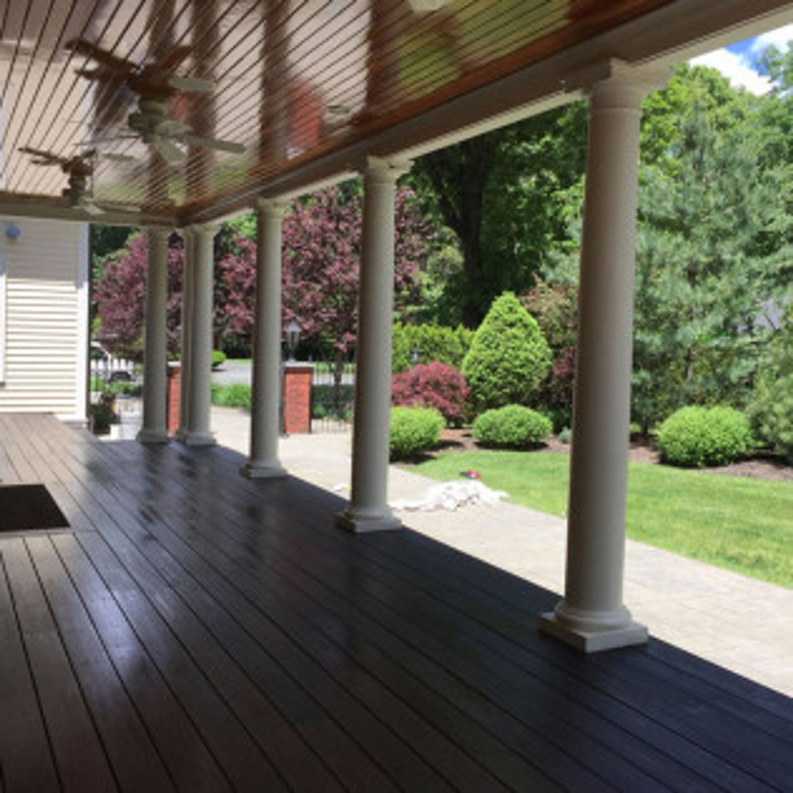 Covered porch with dark wood flooring, white columns, and lush landscaping in the background.