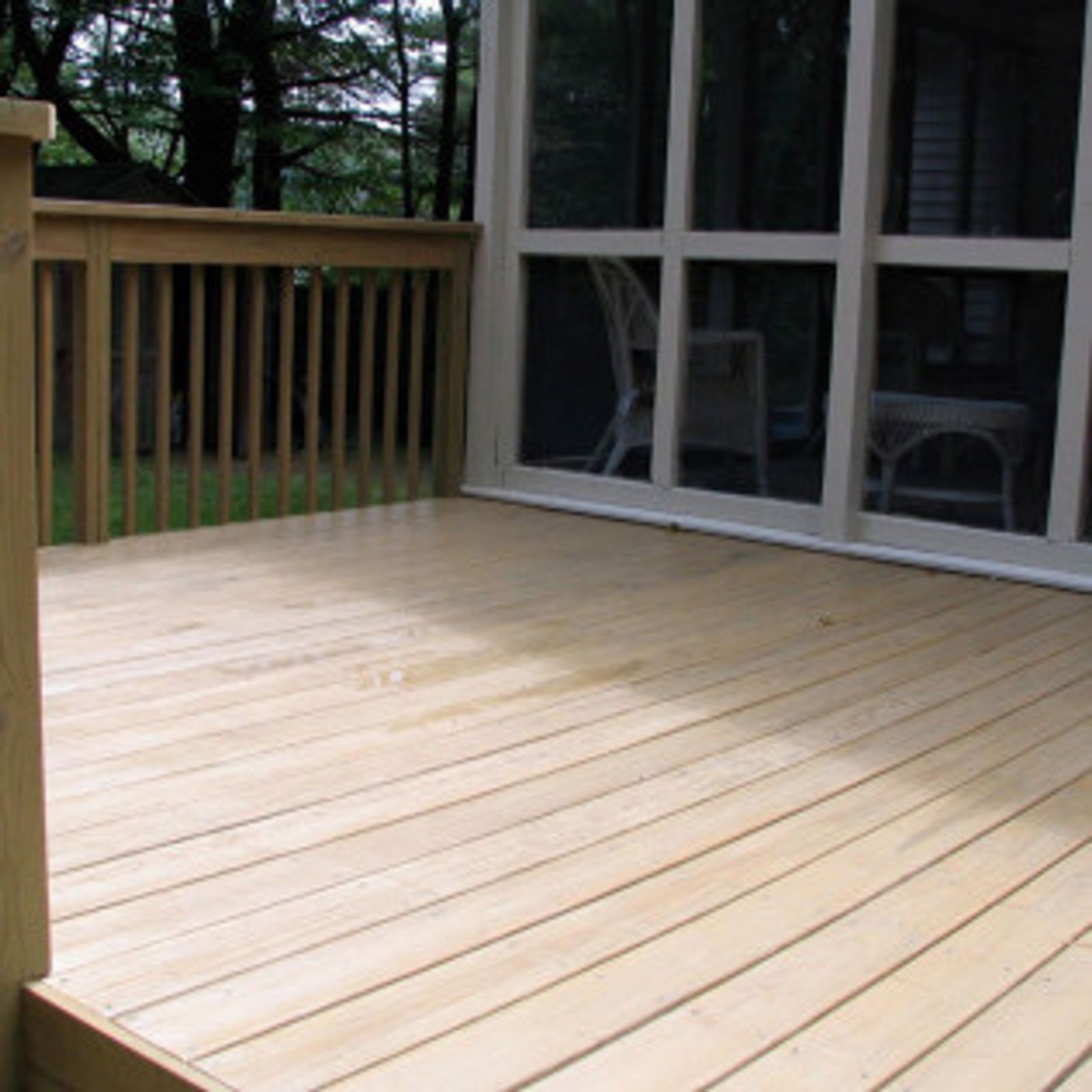 Wooden deck with railing, connected to a screened porch. Trees in background.