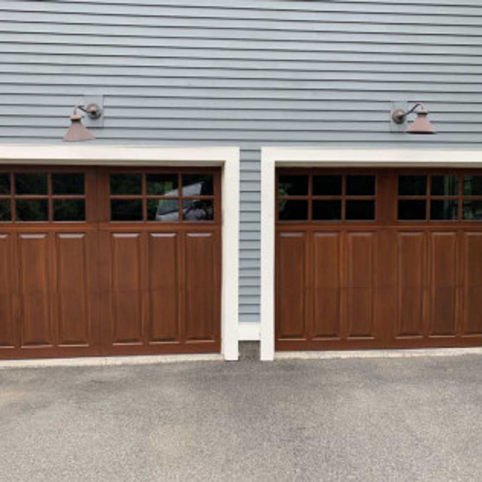Two brown garage doors with glass windows, white trim, and copper lights, set against gray siding.