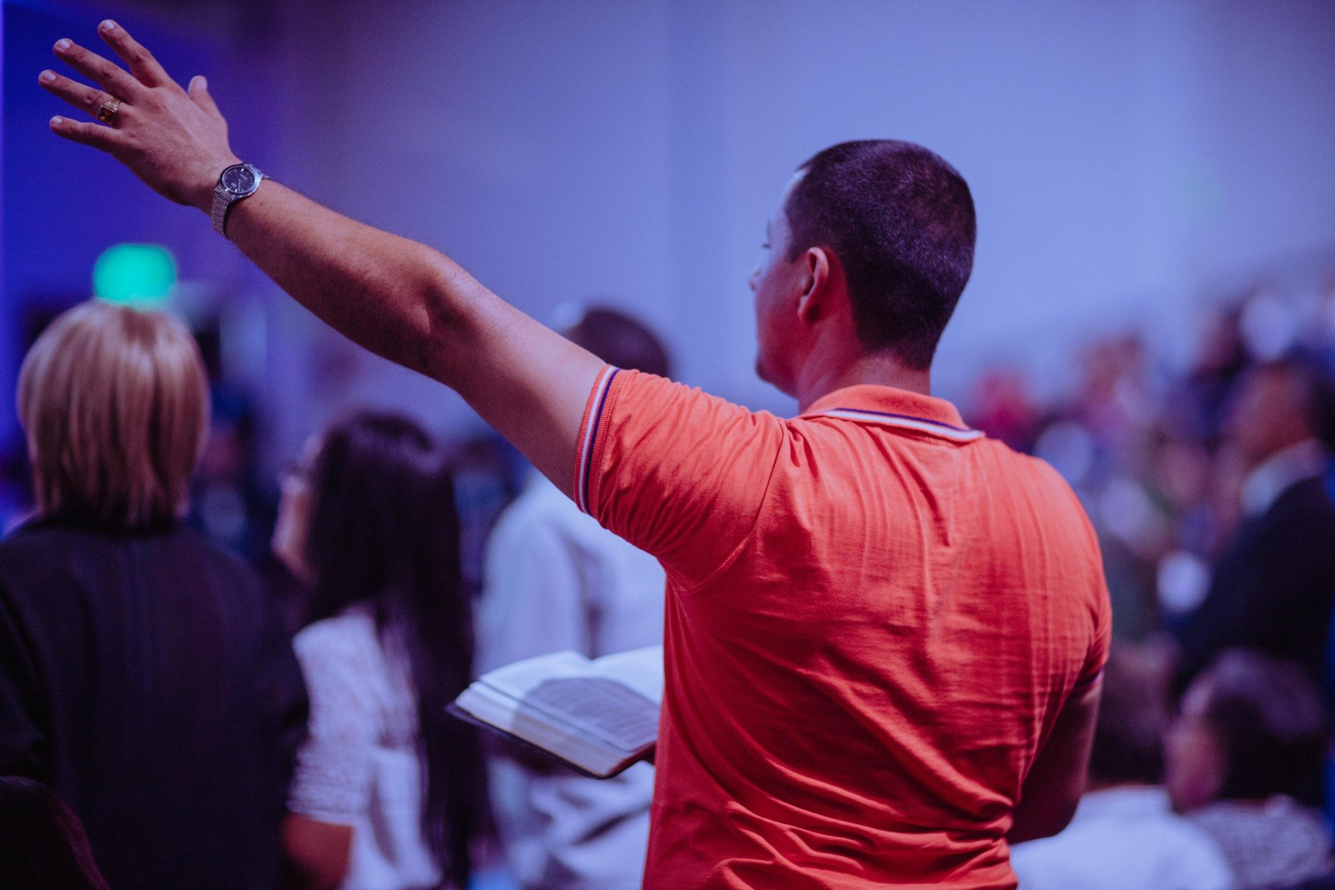 A man in an orange shirt is holding a bible in front of a crowd of people.