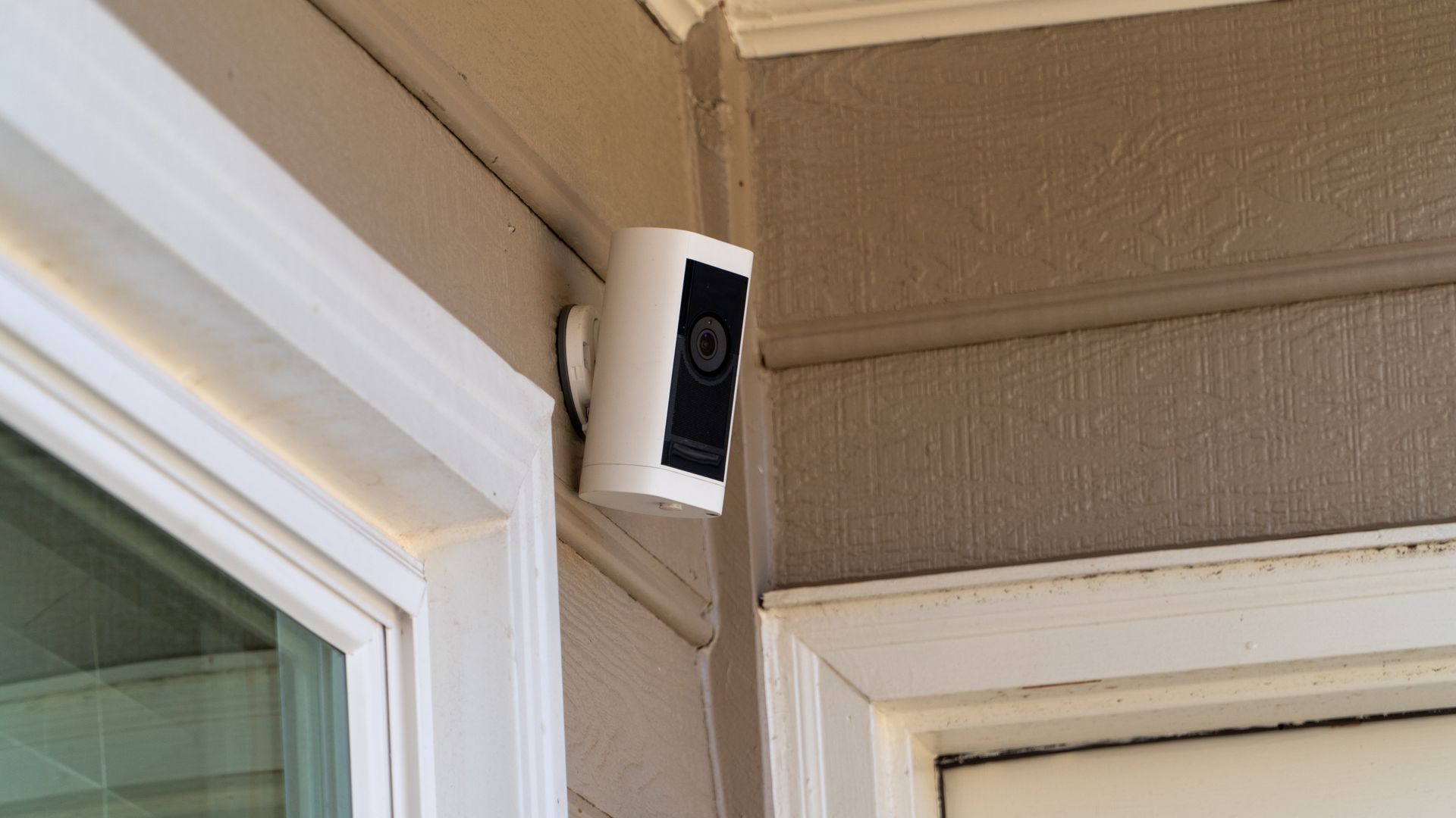 A white Ring security camera mounted on the corner of a beige house exterior near a window and door frame.