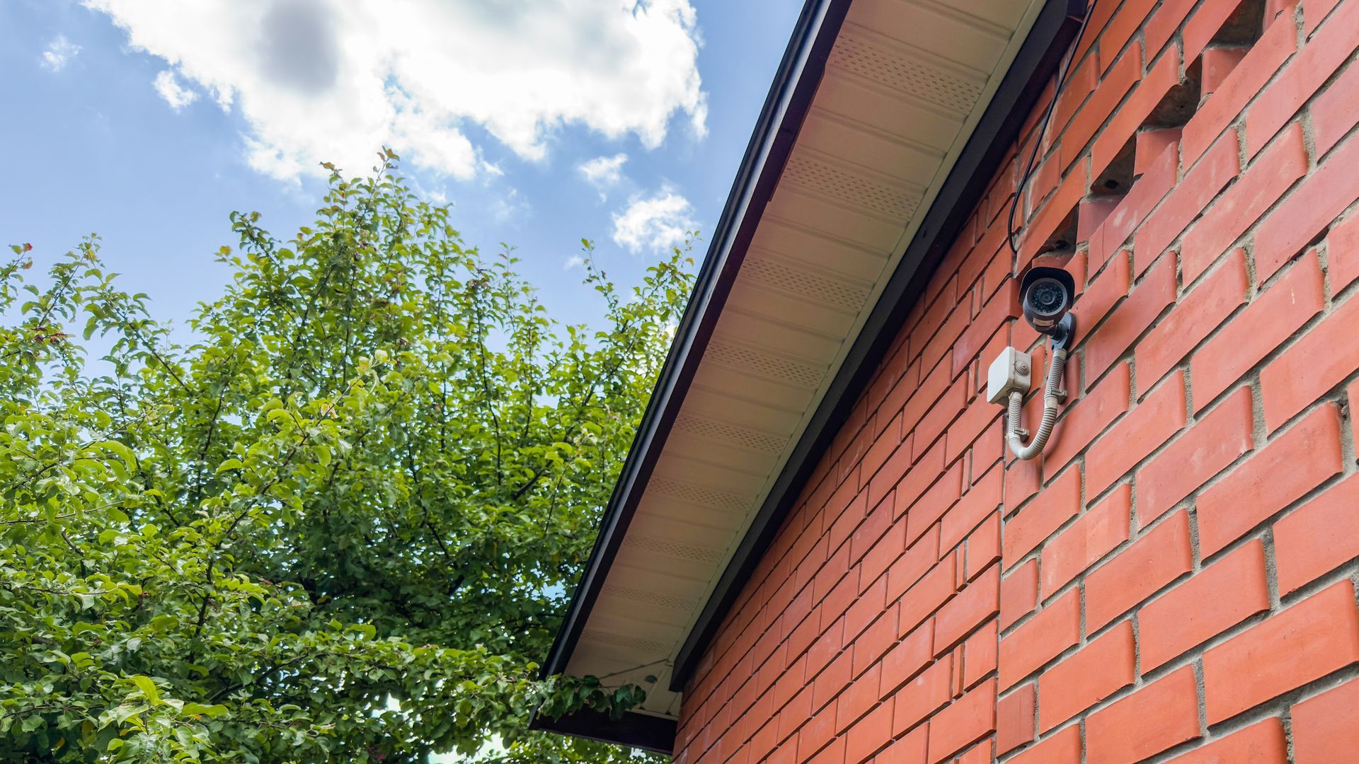 Red brick house with roof camera under blue sky and leafy tree.