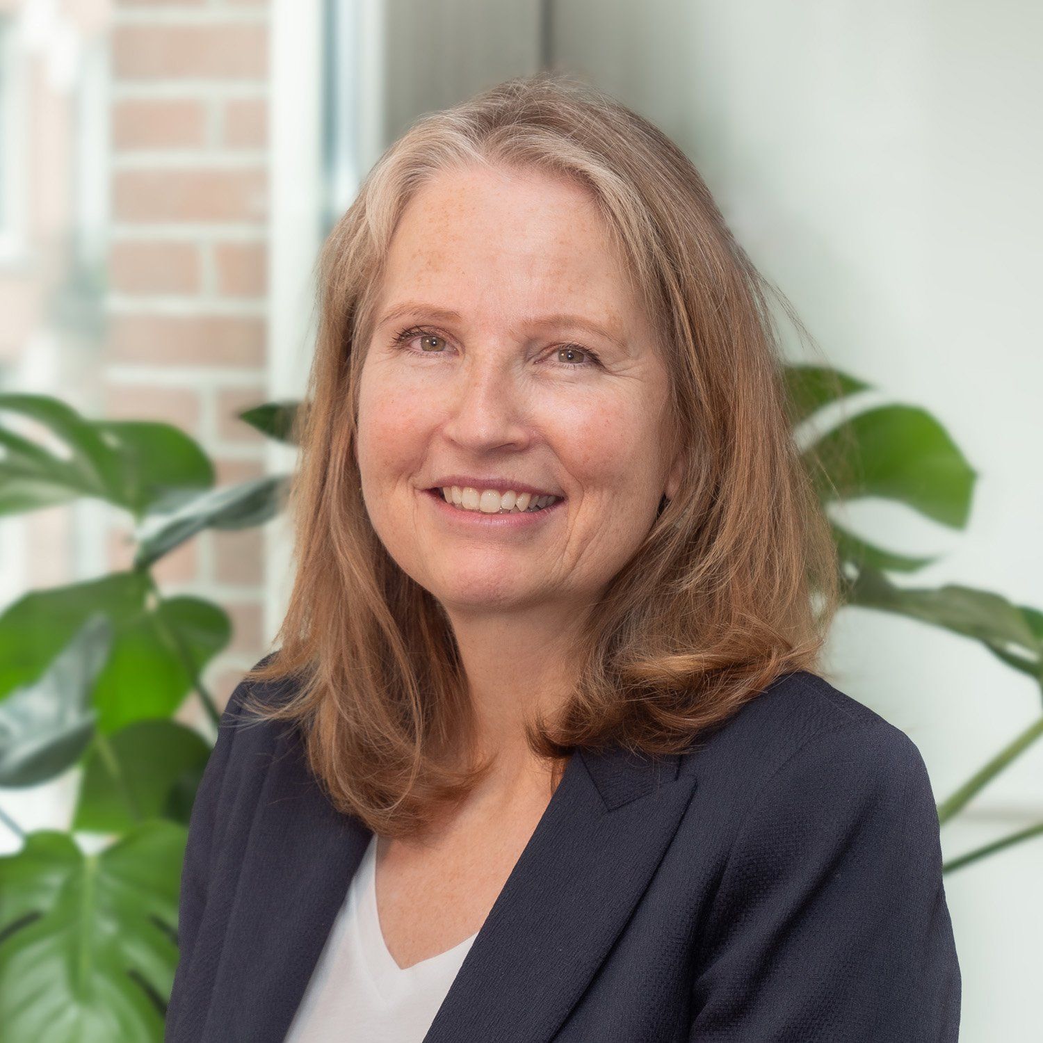 Een persoon met schouderlang haar, gekleed in een donkere blazer over een wit overhemd, lacht voor een groene, bladerrijke plant.