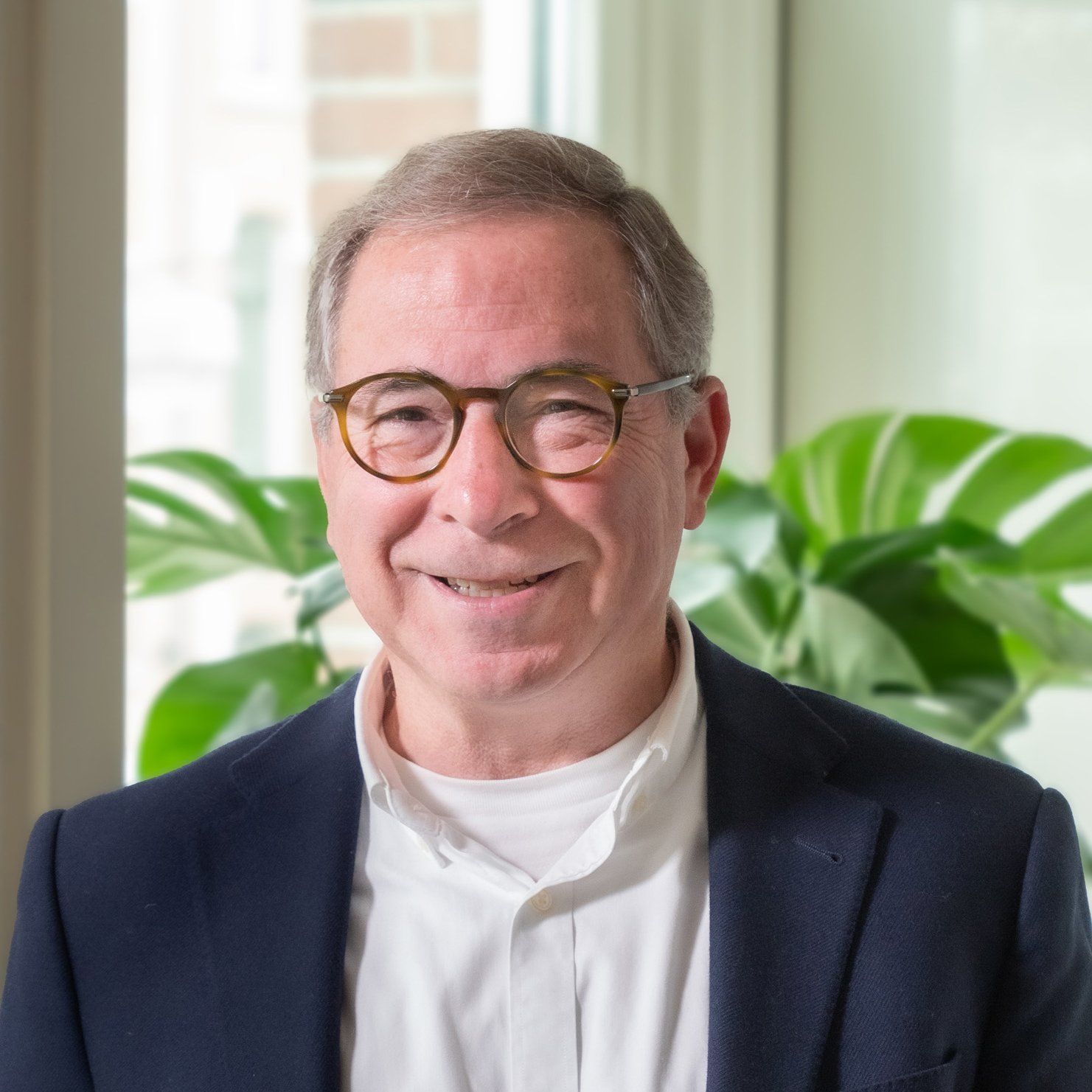 Een glimlachend persoon met een bril, gekleed in een wit overhemd en een donkerblauwe blazer, staat voor een groene plant.