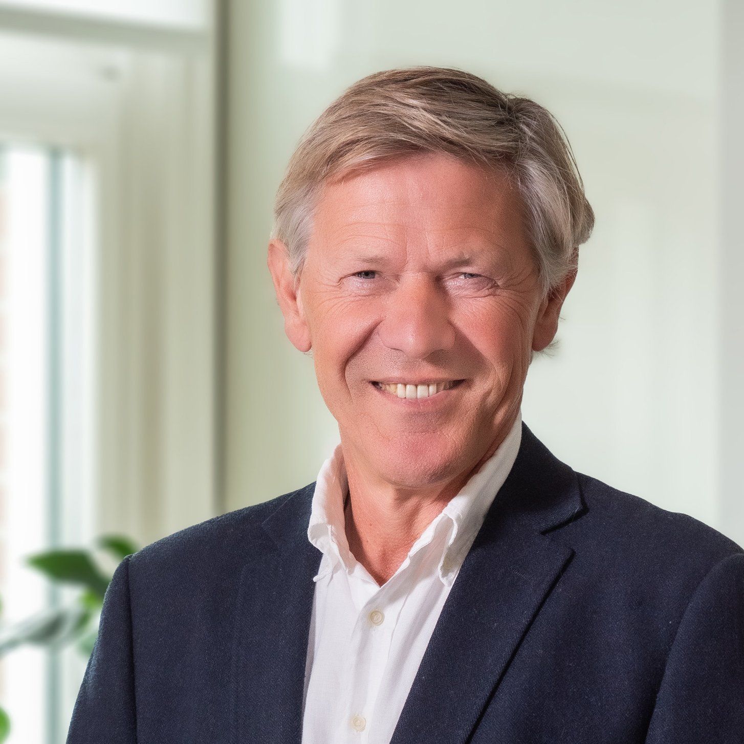 Een glimlachende persoon met lichtbruin haar draagt een donkerblauwe blazer en een wit overhemd in een sfeervol verlichte kantooromgeving.