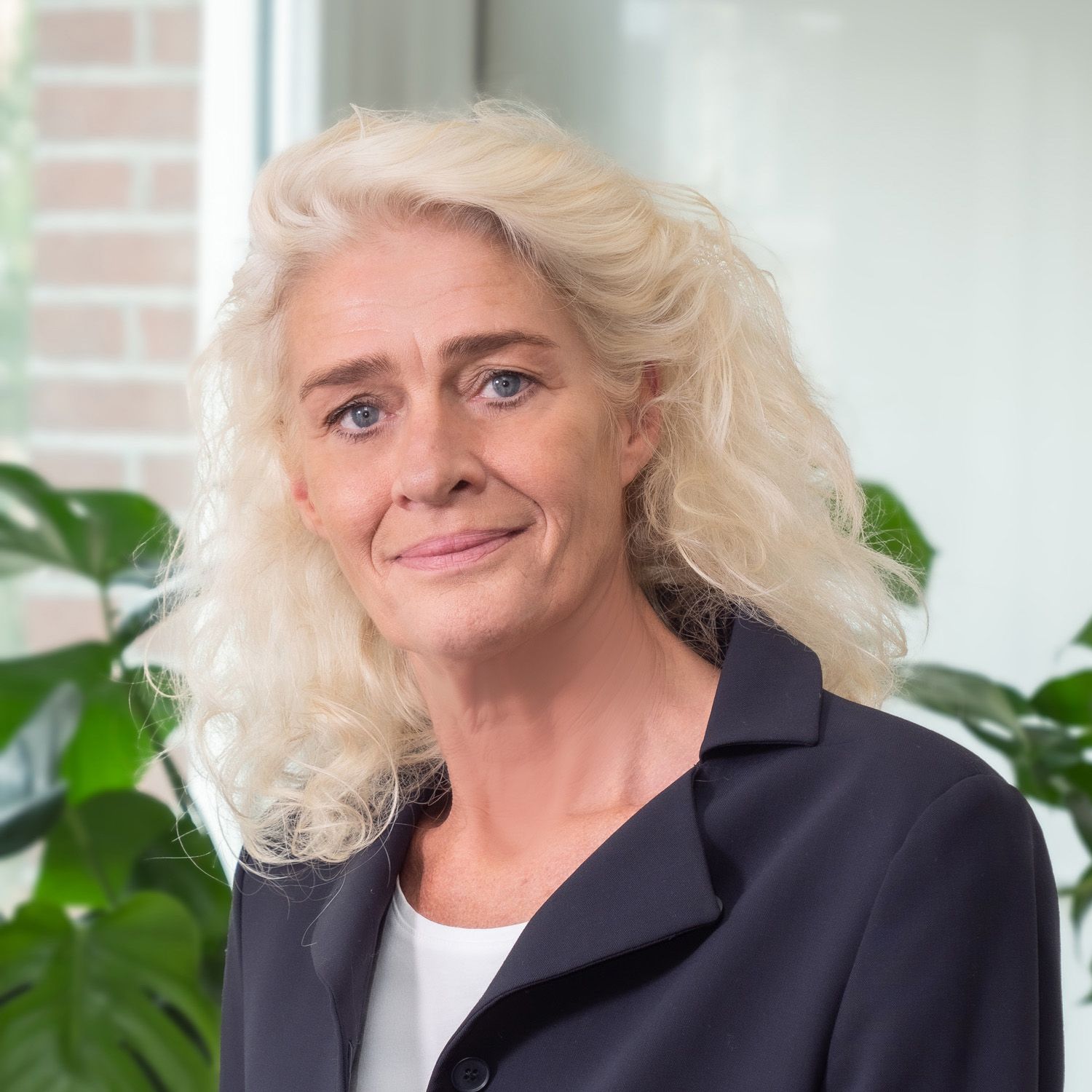 Een professioneel portret van een persoon met blond, golvend haar, gekleed in een donkerblauwe blazer, poserend voor kasplanten.