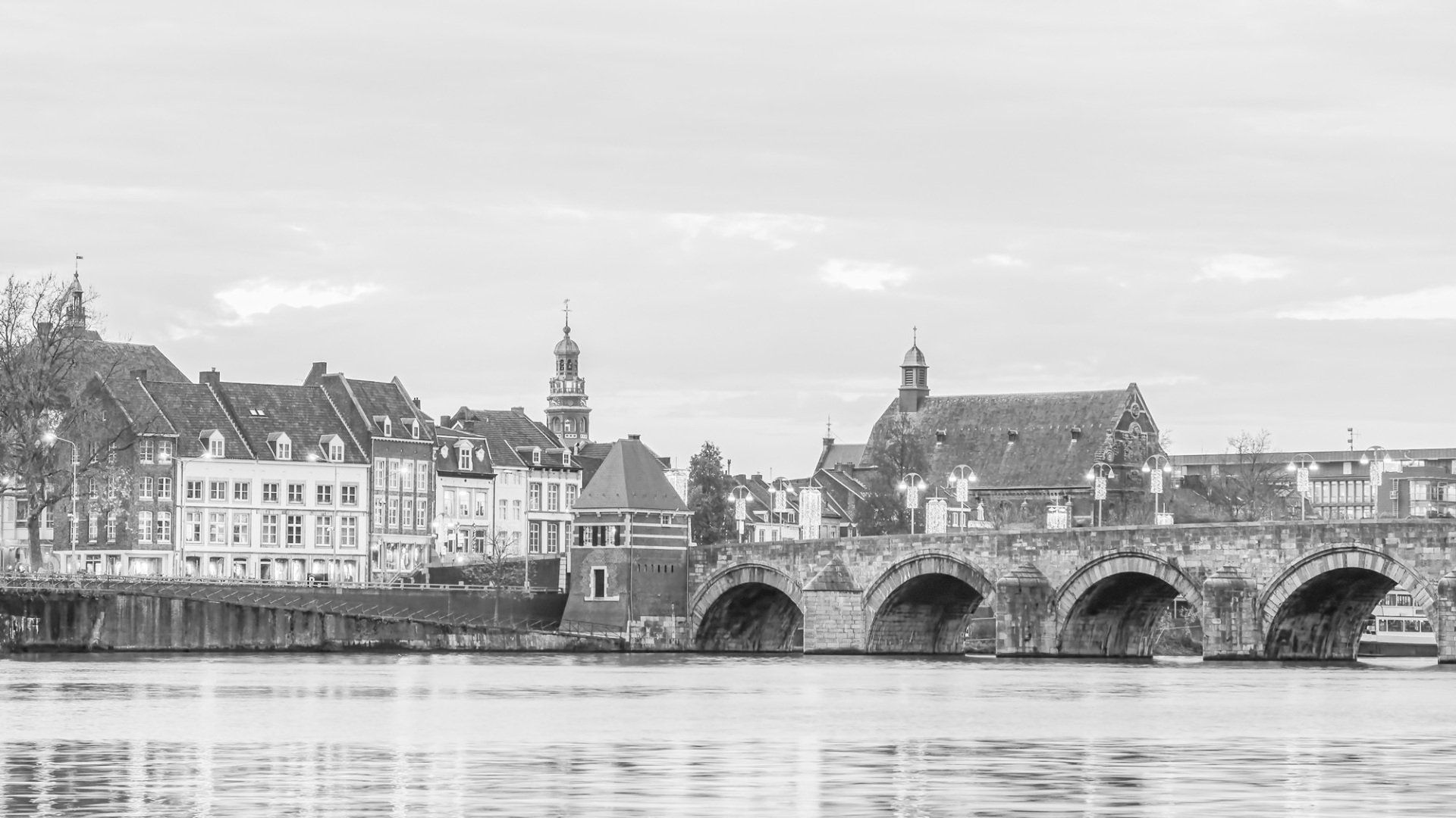 Een zwart-witfoto van een stenen boogbrug over een rivier, met historische gebouwen en kerktorens op de achtergrond.