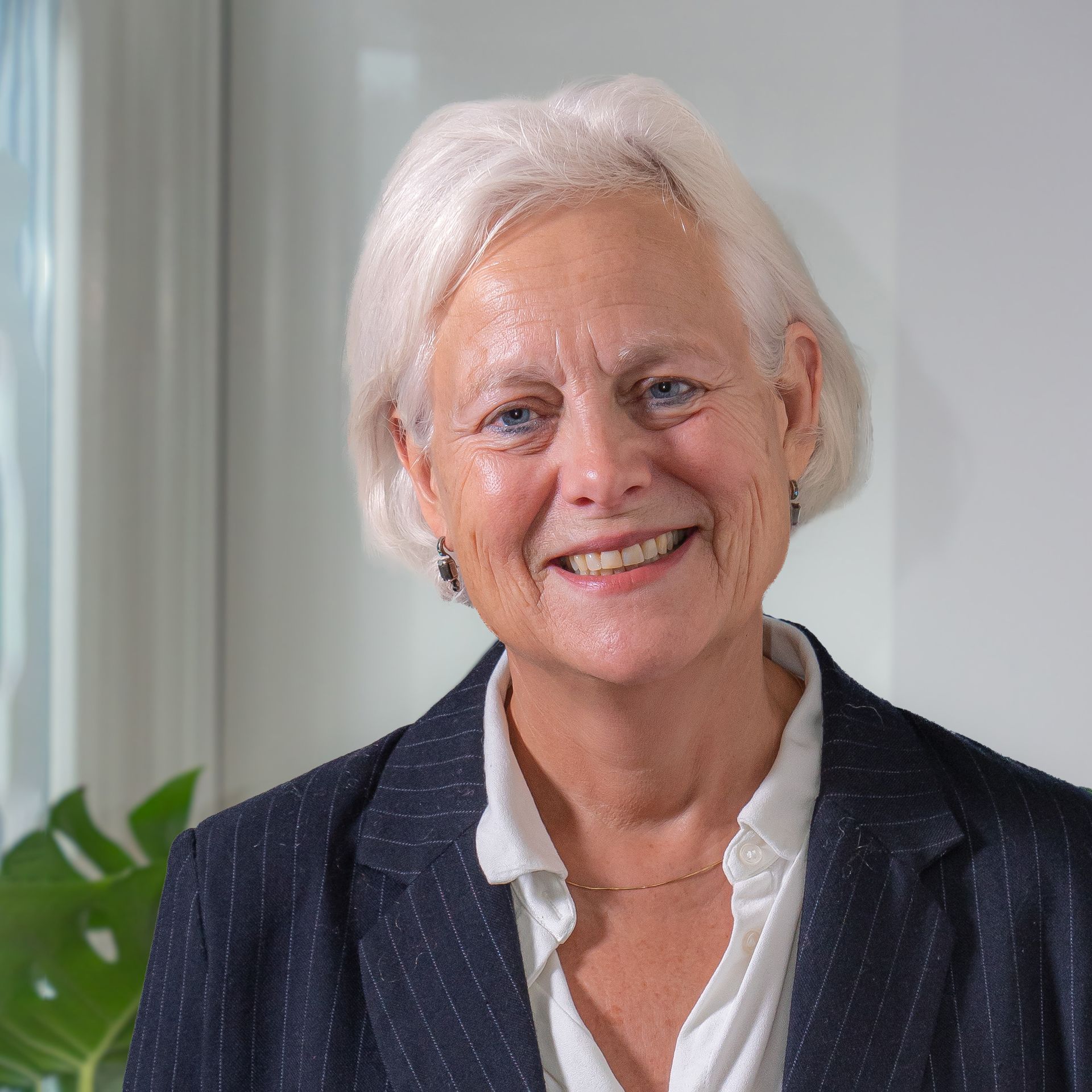 Een lachende persoon met wit haar, gekleed in een wit overhemd en een colbert met krijtstrepen, poseerde in een lichte kantoorruimte.