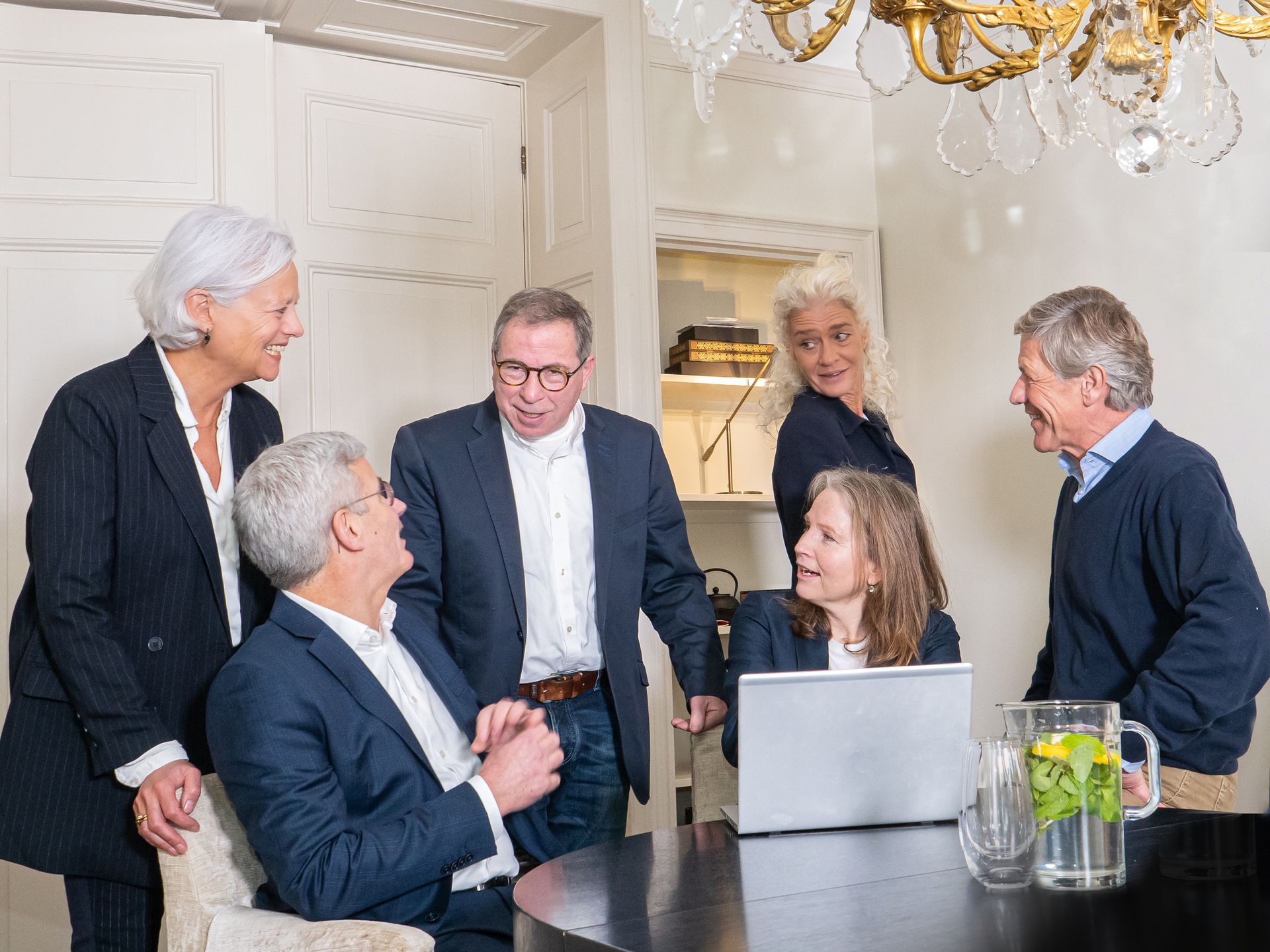 Een groep professionals in zakelijke kleding zit rond een laptop aan een tafel en bespreekt werk in een kantooromgeving.