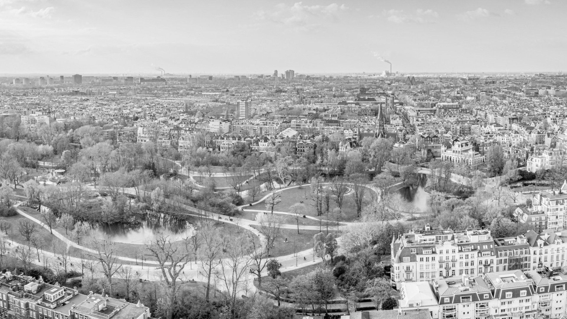 Een zwart-wit luchtfoto van een groot park met een meer, omgeven door stadsgebouwen en dichte bomen.